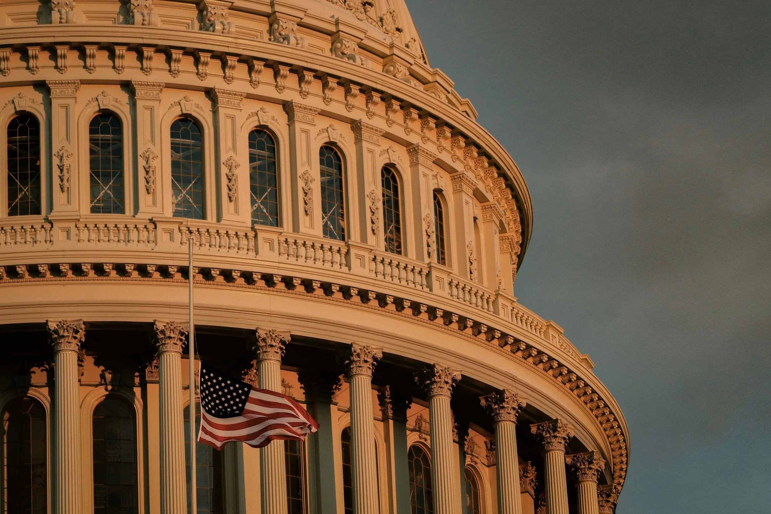 The United States Capitol building illuminated by the golden light of sunset, with architectural details and decorative columns visible, and an American flag on display.