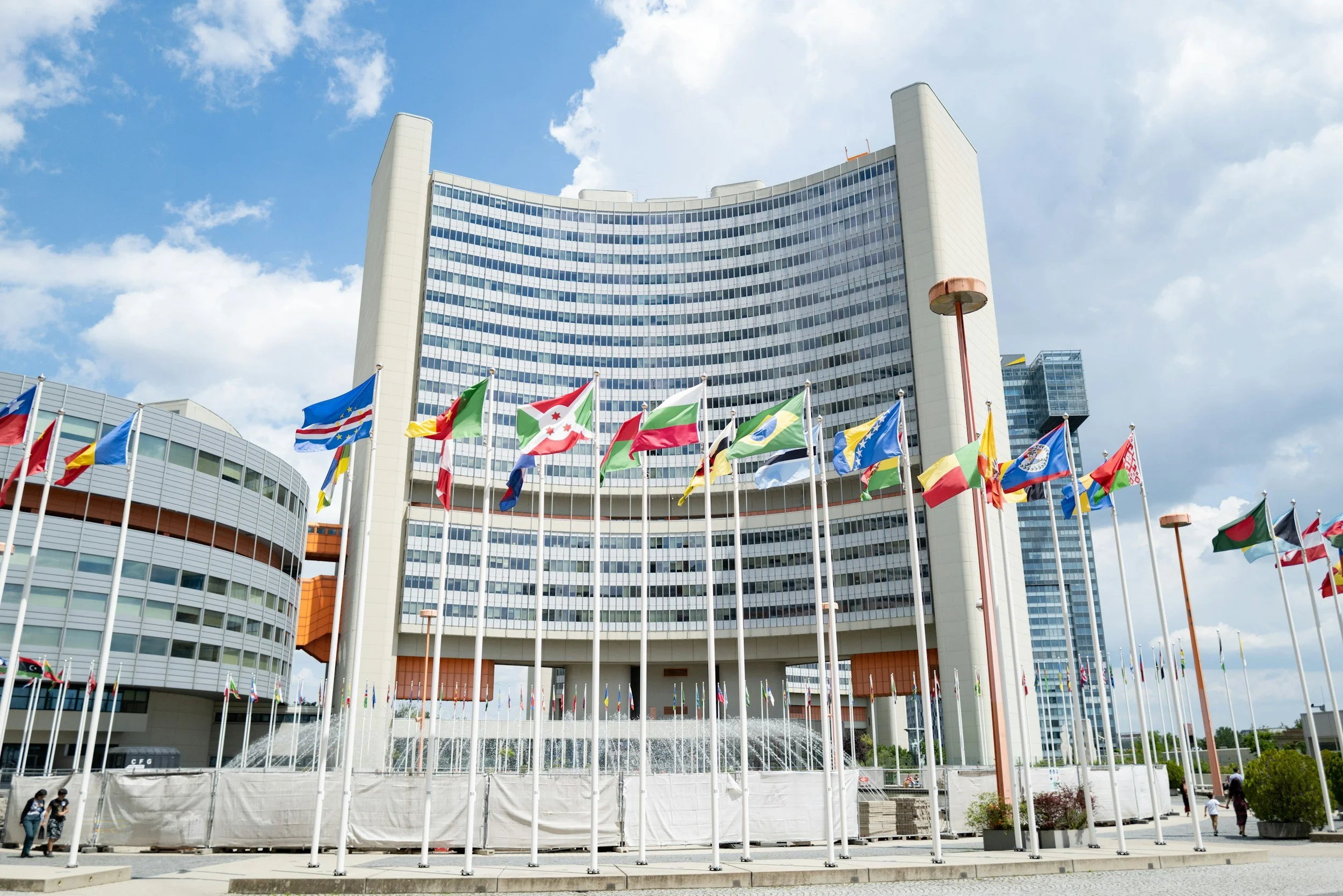 Modern multistory government or office building with a curved facade, displaying multiple flags from different countries in front, under a partly cloudy sky.