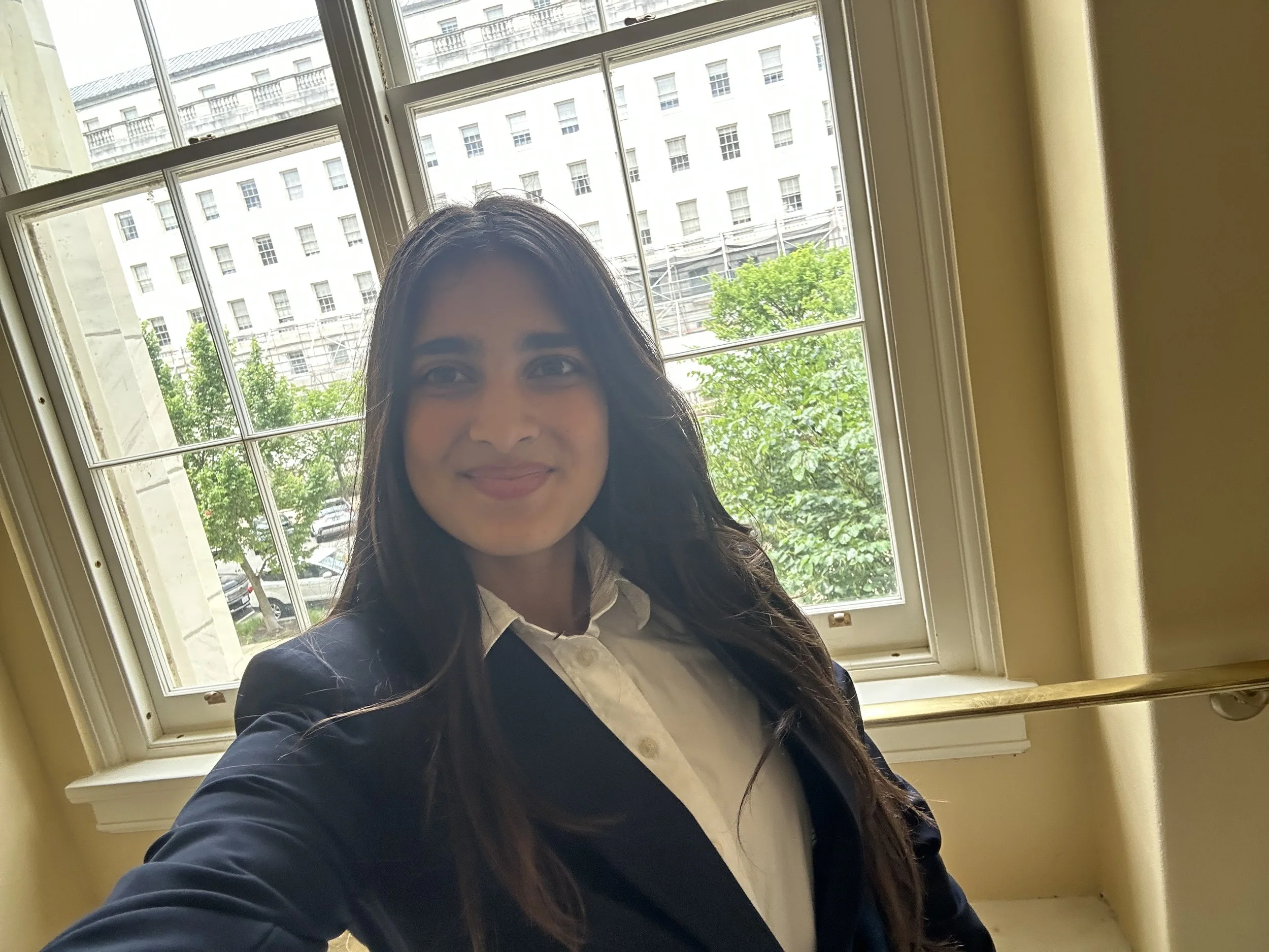 A woman taking a selfie near a large window with a cityscape view, including a white building and trees outside.