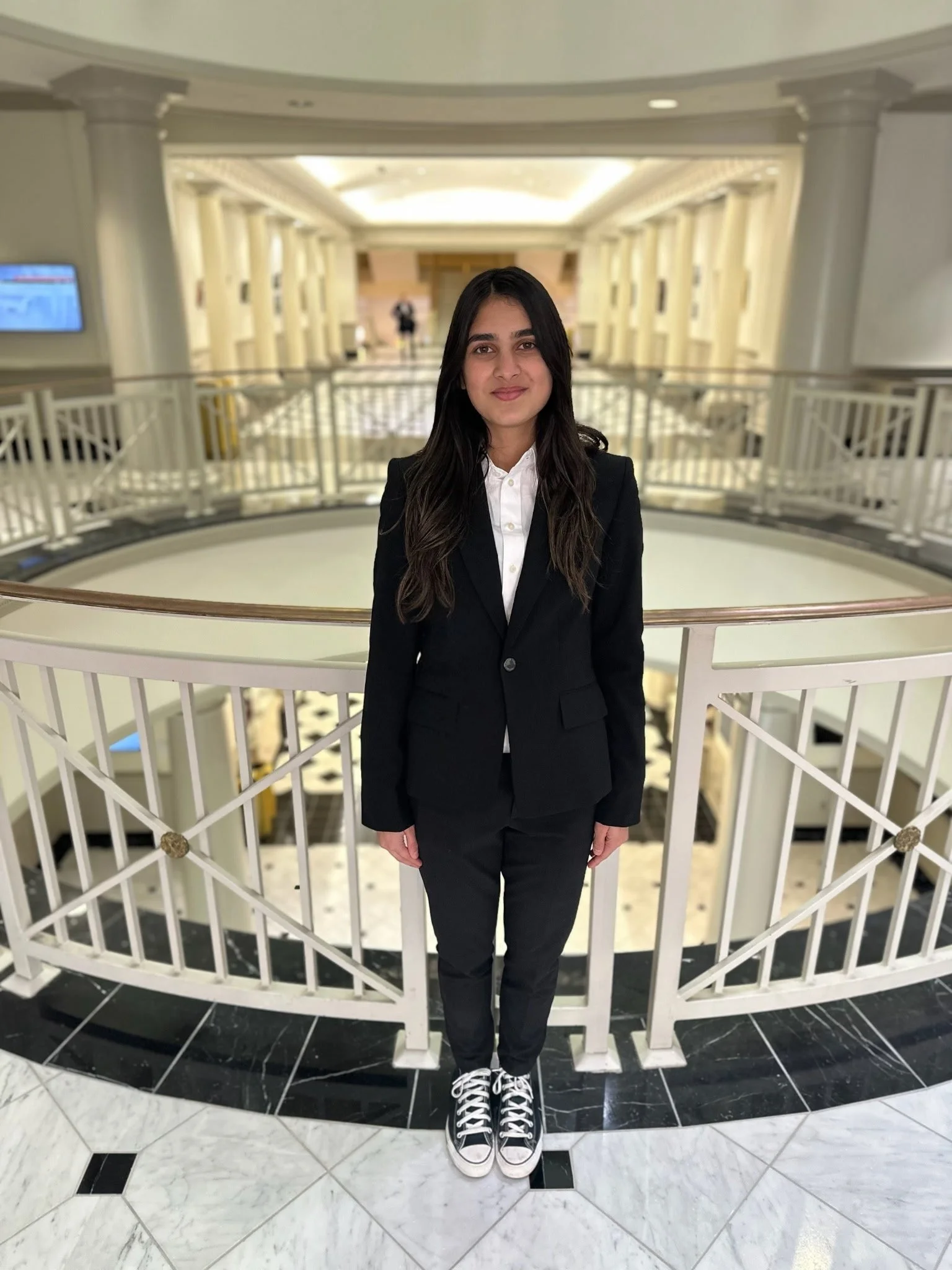 Young woman in black suit and white shirt standing in front of a white round railing inside a luxurious building with marble flooring and warm lighting.