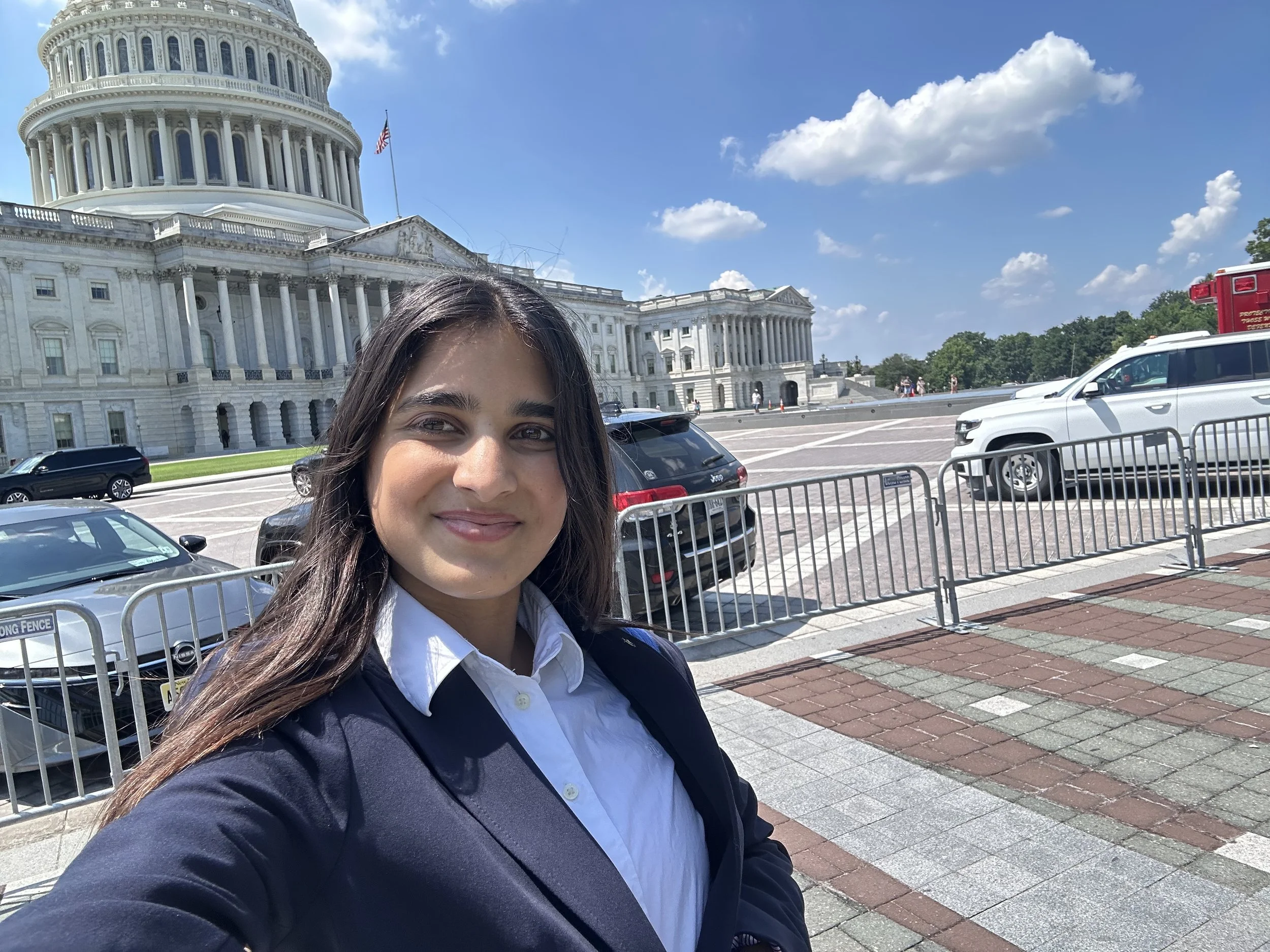 A woman smiling in front of the United States Capitol building on a sunny day, with parked cars and a fence in the foreground.
