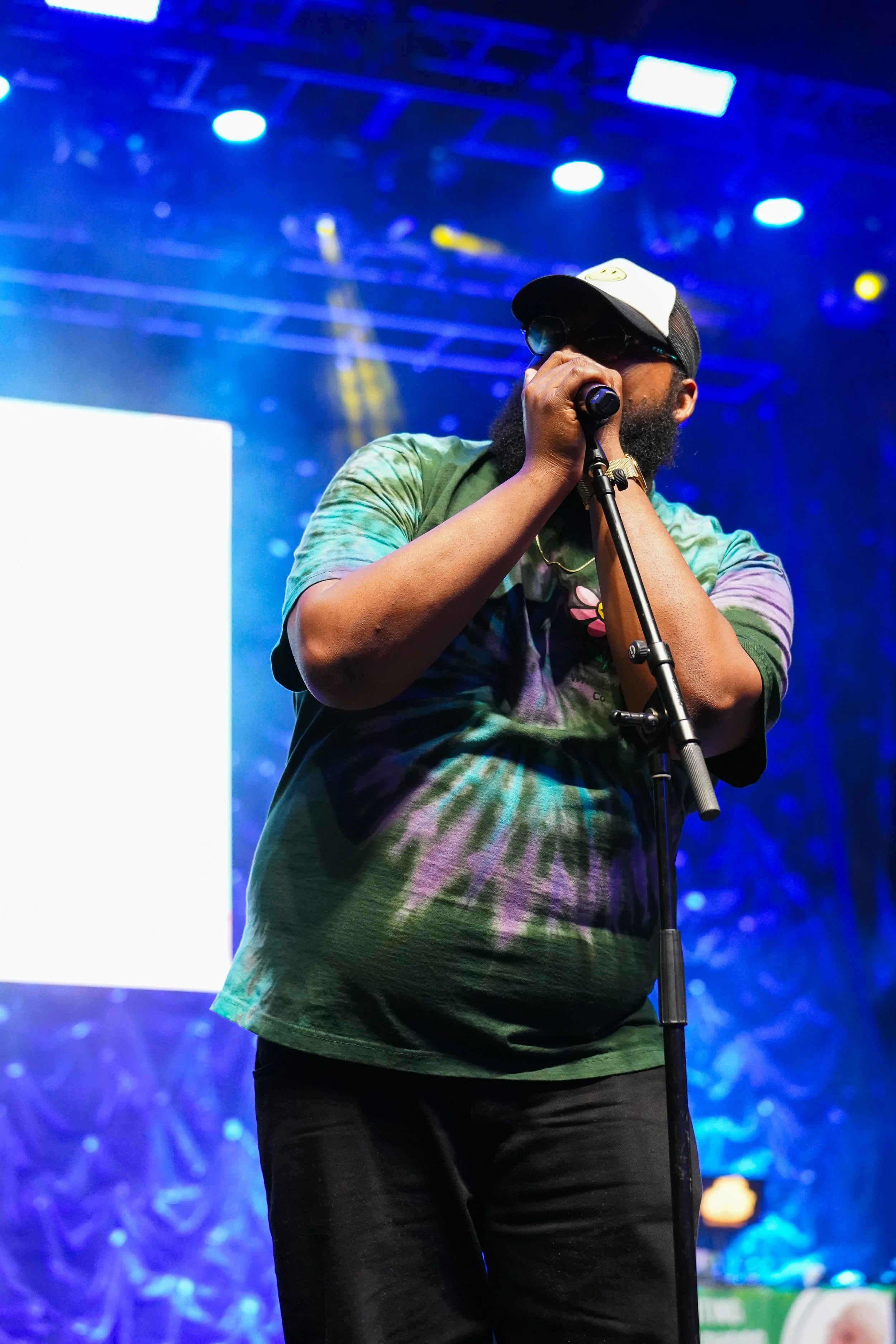 A man wearing a rainbow tie-dye shirt, black pants, hat, sunglasses, and a face mask is performing on stage with a microphone in hand. There are bright stage lights and a large screen in the background.
