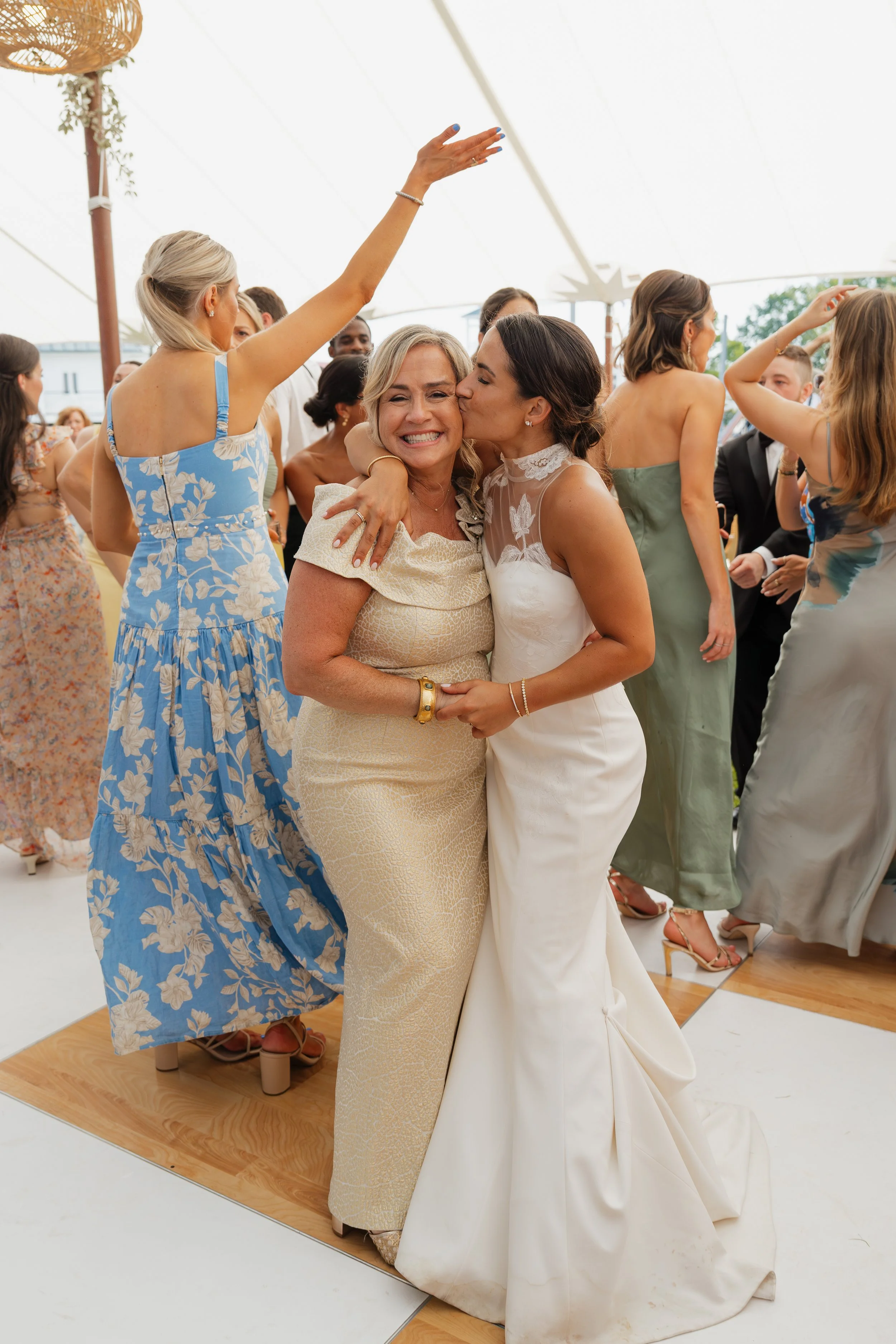 A woman in a white wedding dress and a woman in a light-colored dress share a loving moment, with the woman in the wedding dress kissing the older woman on the cheek during a wedding celebration. They are surrounded by guests dancing and celebrating under a white tent.