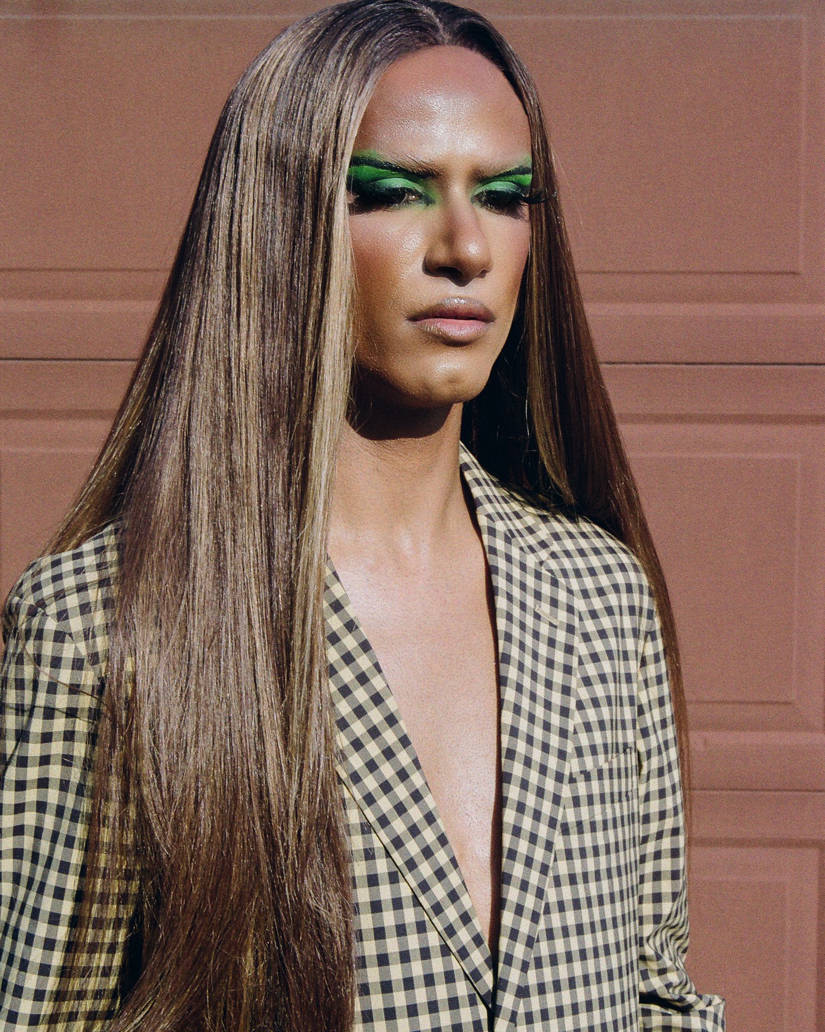Image of a drag artist wearing a checkered blazer against a brown wall on film