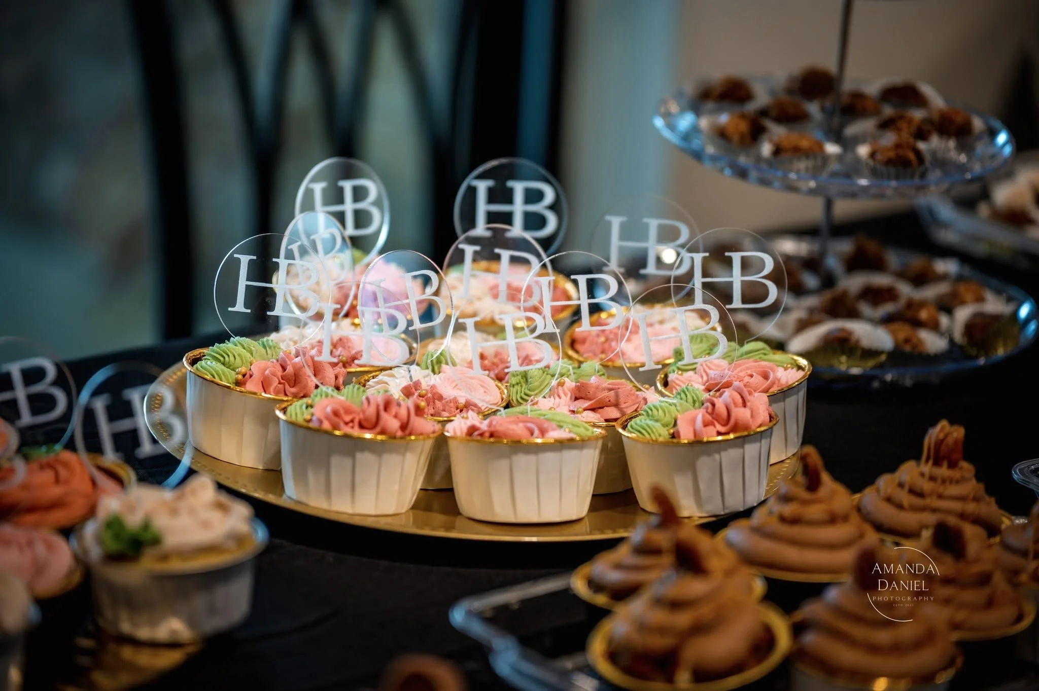 A dessert table with cupcakes decorated with pink, green, and white frosting, topped with 'HB' themed toppers, and a tiered tray of cookies in the background.
