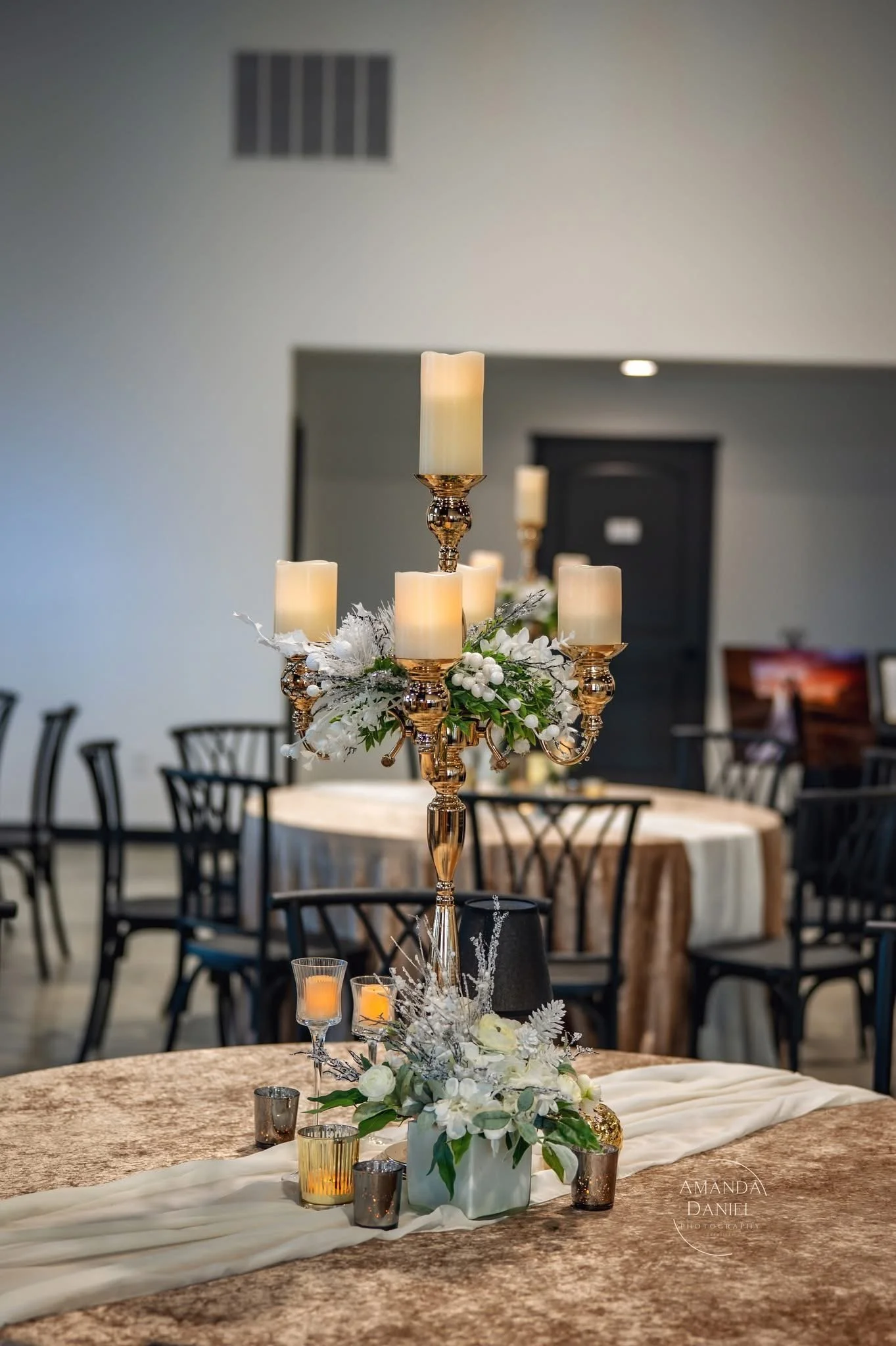 Elegant table centerpiece with a gold candelabrum holding five candles, surrounded by white flowers and decorative accents, on a brown tablecloth in a banquet hall.