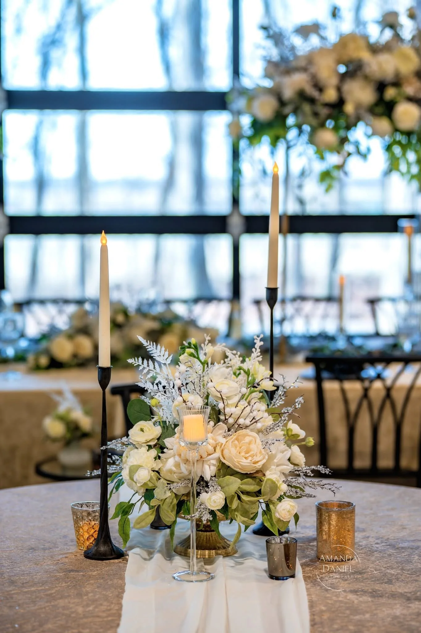 Elegant wedding table centerpiece with white flowers, candles, and metallic candle holders in a well-lit venue.