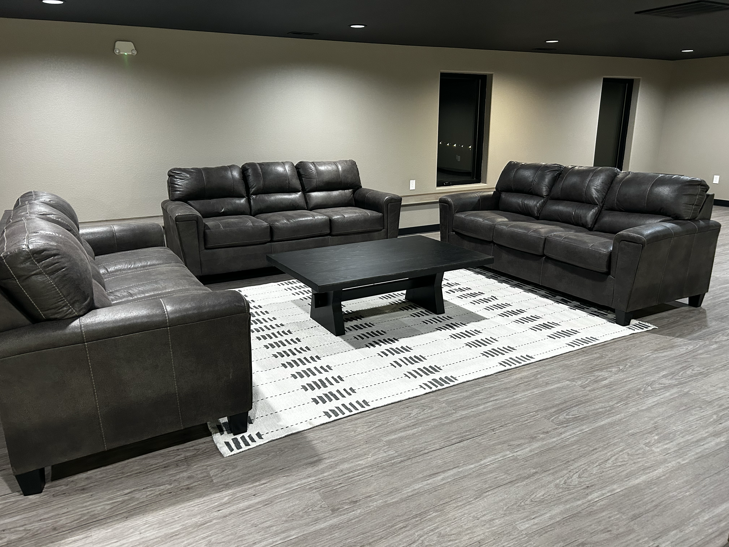 Three black leather sofas arranged around a black coffee table on a white and black patterned rug in a modern basement lounge with beige walls and wood-like flooring.