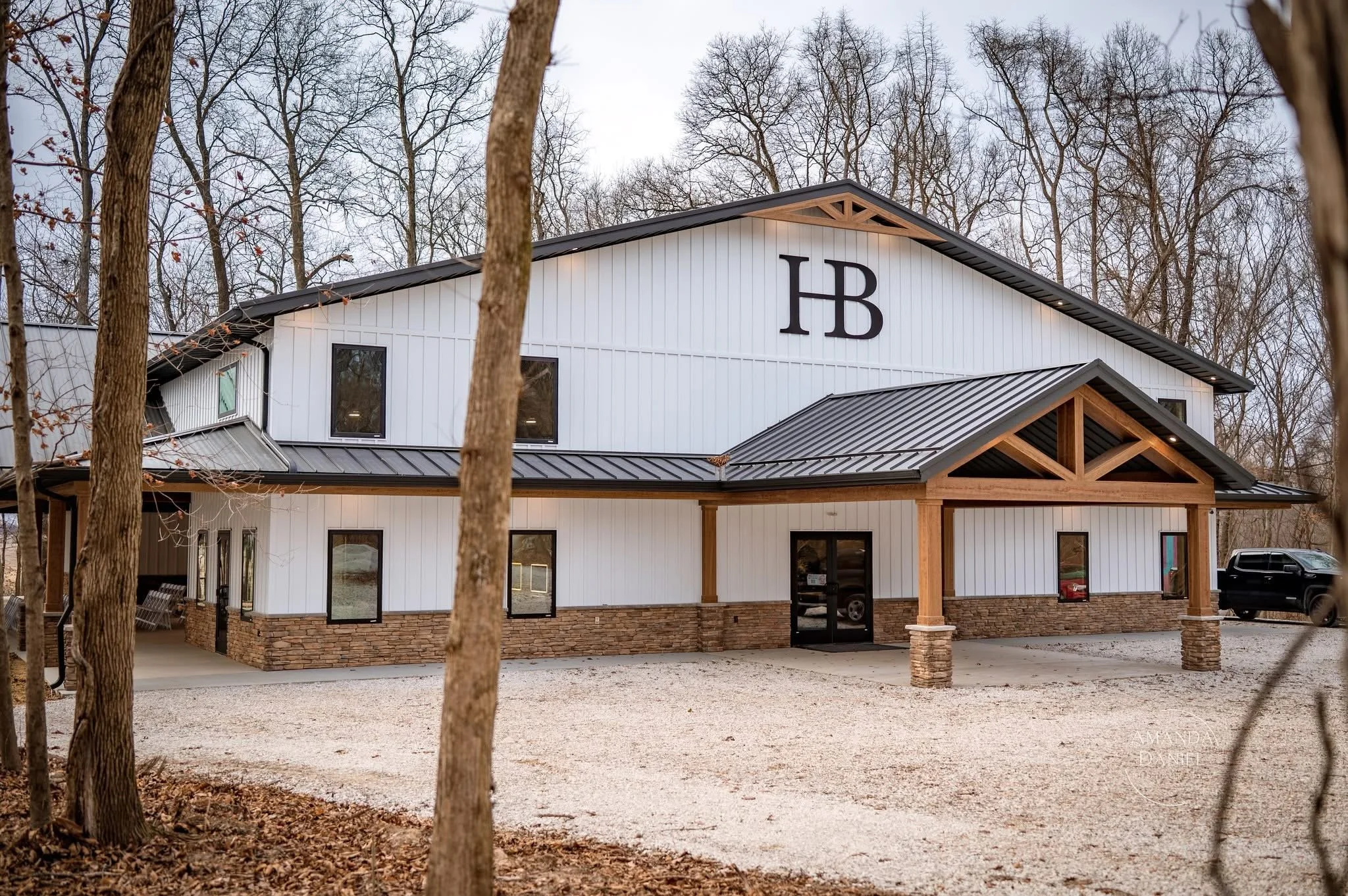 A modern barn-style building with white siding, a black metal roof, and wooden accents, surrounded by leafless trees and a gravel driveway.