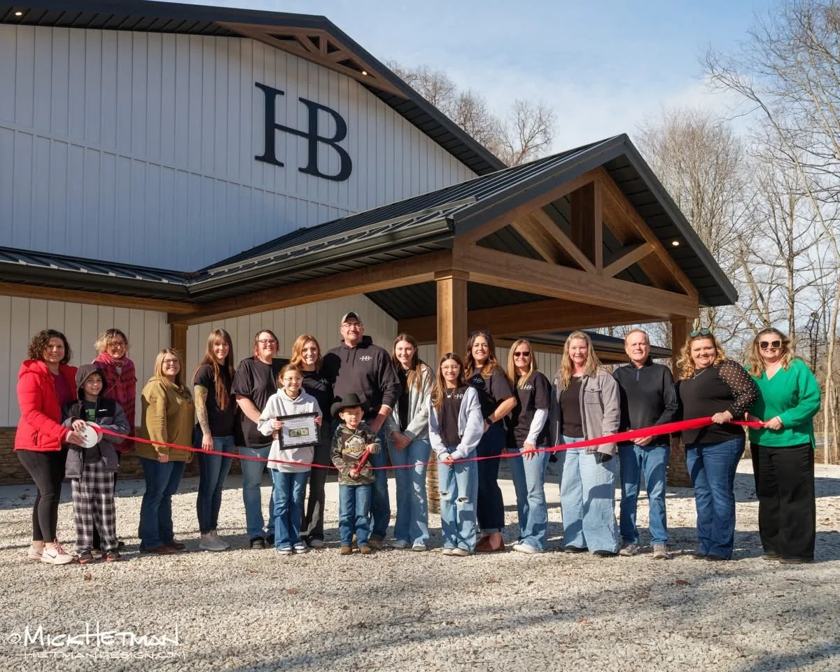 A group of people gathered for a ribbon-cutting ceremony outside a building with a large HB logo on it. The group includes children and adults, some holding the red ribbon and scissors, celebrating the opening of a new facility.
