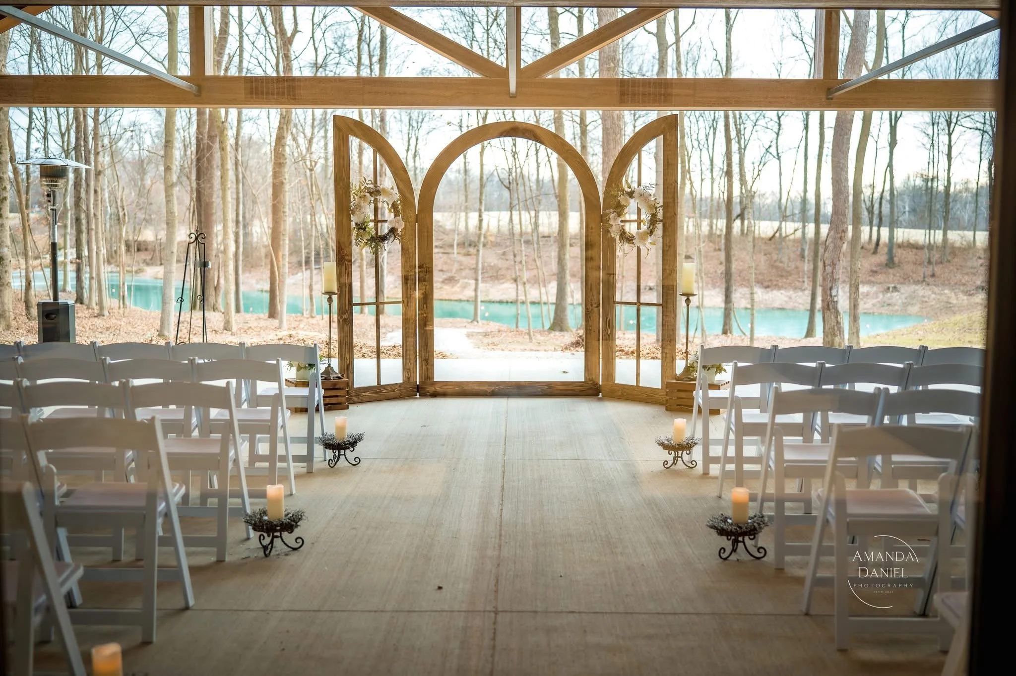 Inside a rustic outdoor wedding venue with white folding chairs arranged in rows, wooden arch with floral decorations, candles, and a view of a lake and trees in the background.