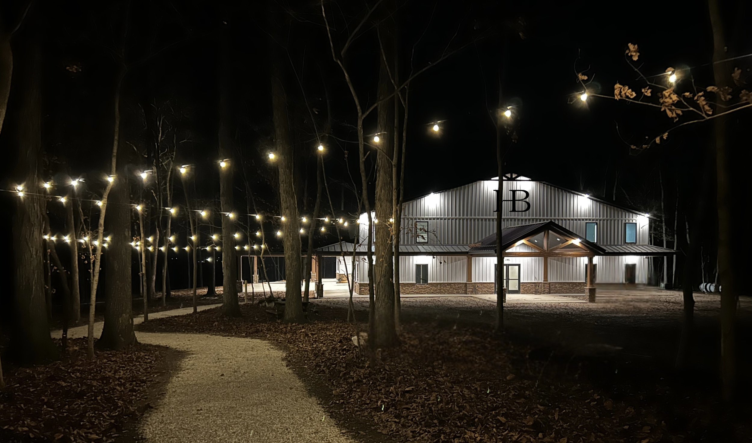 Nighttime scene with string lights hanging above a winding gravel path through a wooded area leading to a large metal barn with a lit entrance and a prominent letter 'HB' on its front.