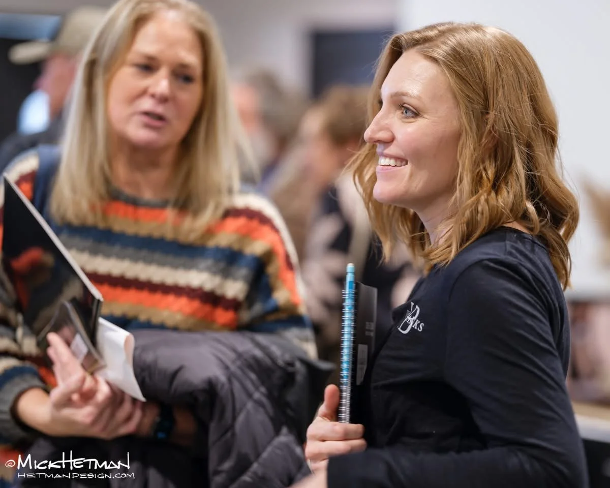 Two women talking at a busy indoor event, one smiling and holding notebooks and pens, the other speaking with a serious expression.