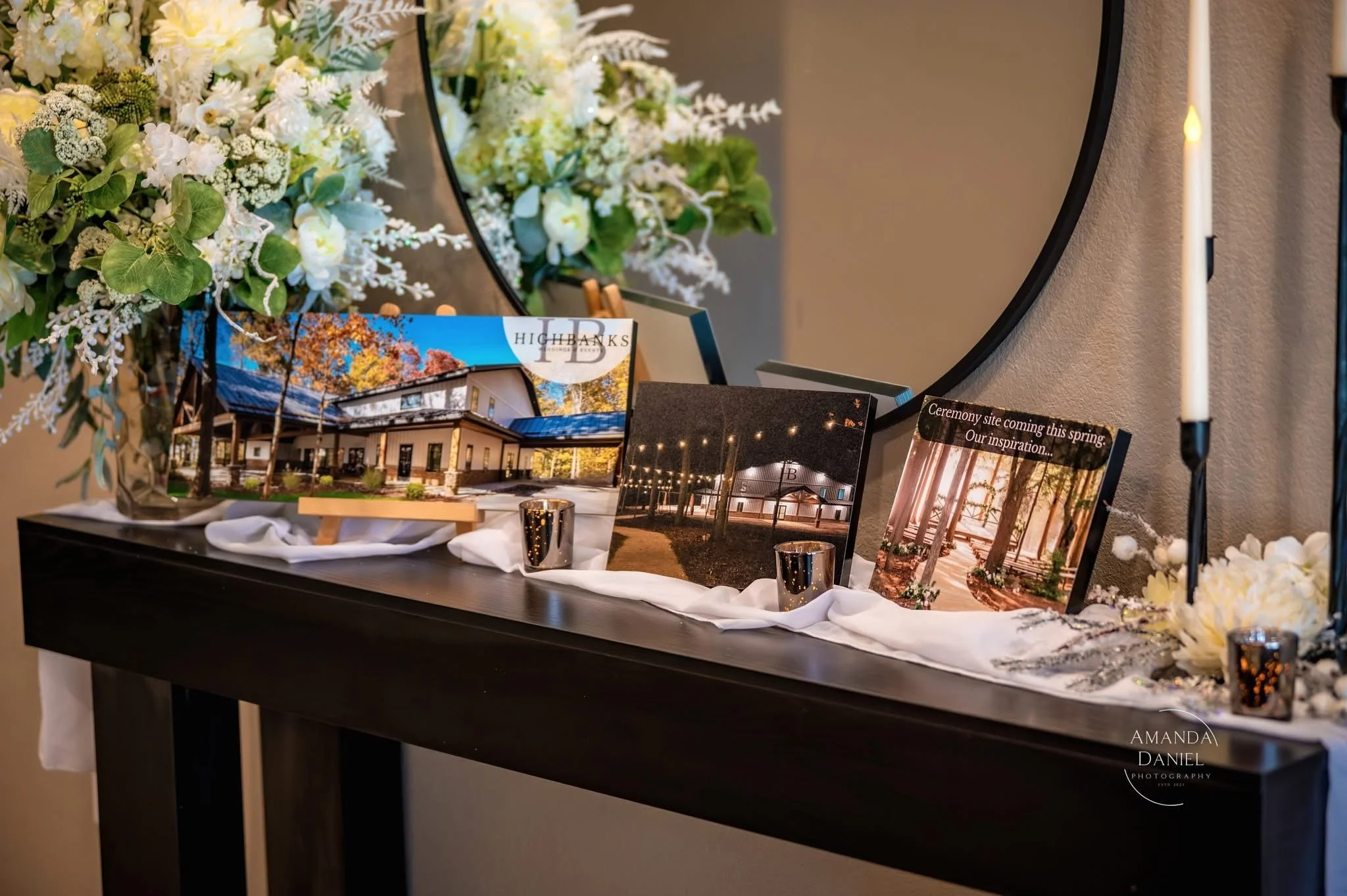 Display table with photographs of a building, a decorated event site, and a ceremony setup, surrounded by floral arrangements, candles, and a mirror.