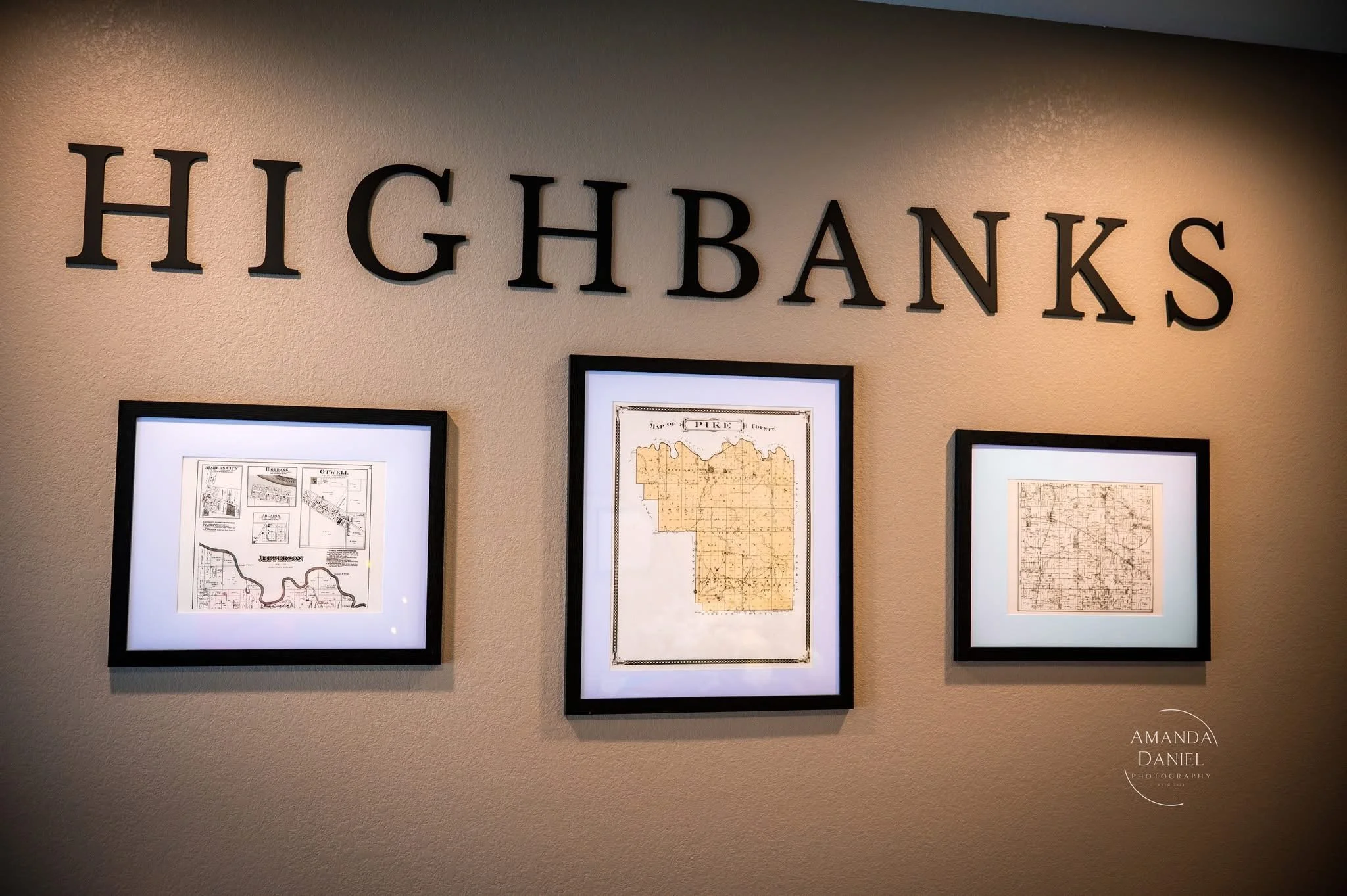 Wall with black letters spelling 'HIGHBANKS' and three framed maps or documents displayed beneath.