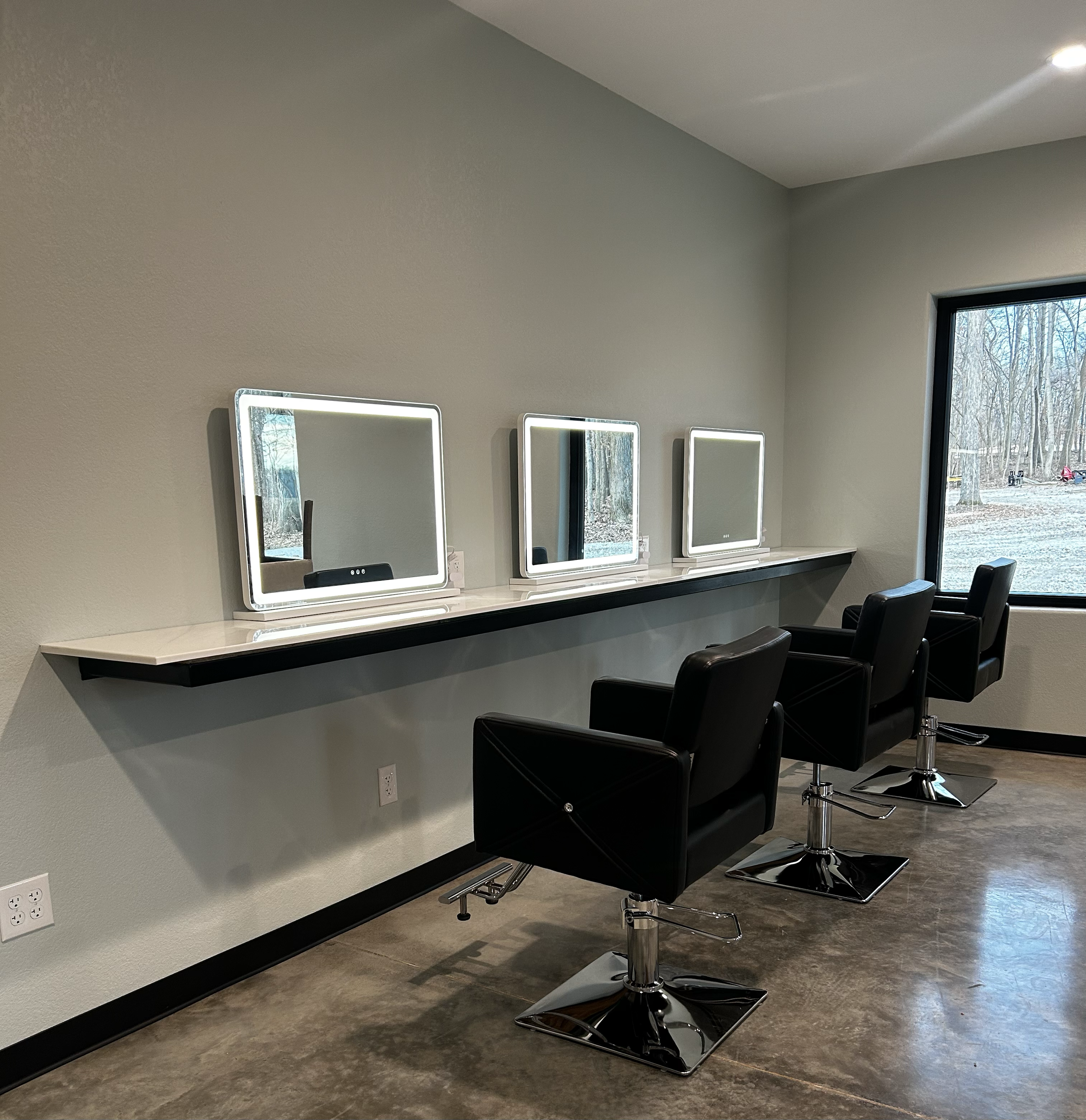 Beauty salon with three illuminated mirrors and black styling chairs near a window showing an outdoor winter scene.