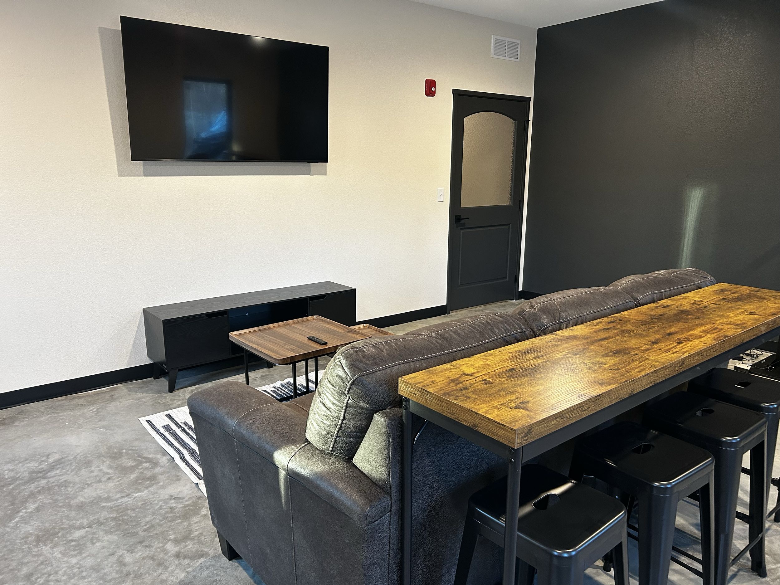 Modern living room with a large wall-mounted TV, a black TV stand, a gray sofa, a wooden coffee table, a striped rug, a long wooden dining table with black stools, dark gray accent wall, and a door.