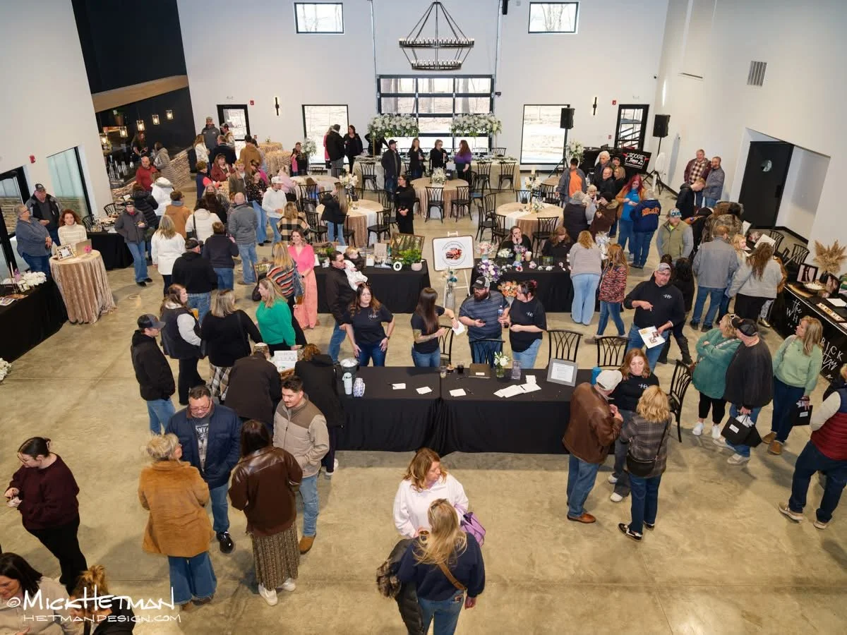 A large indoor event with many people walking around and gathered around tables, some looking at displays, in a spacious hall with high windows and modern decor.