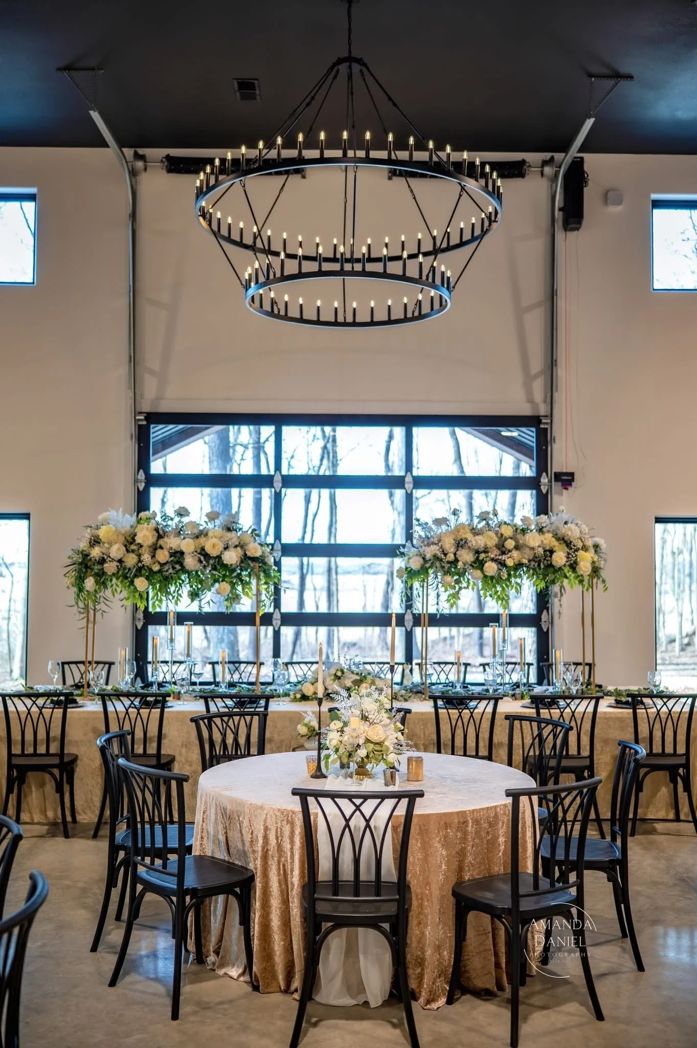 Elegant event space with round table covered in a gold or beige tablecloth, surrounded by black chairs, decorated with white floral centerpieces, tall floral arrangements, and candles. Large windows and an industrial-style chandelier are visible in the background.