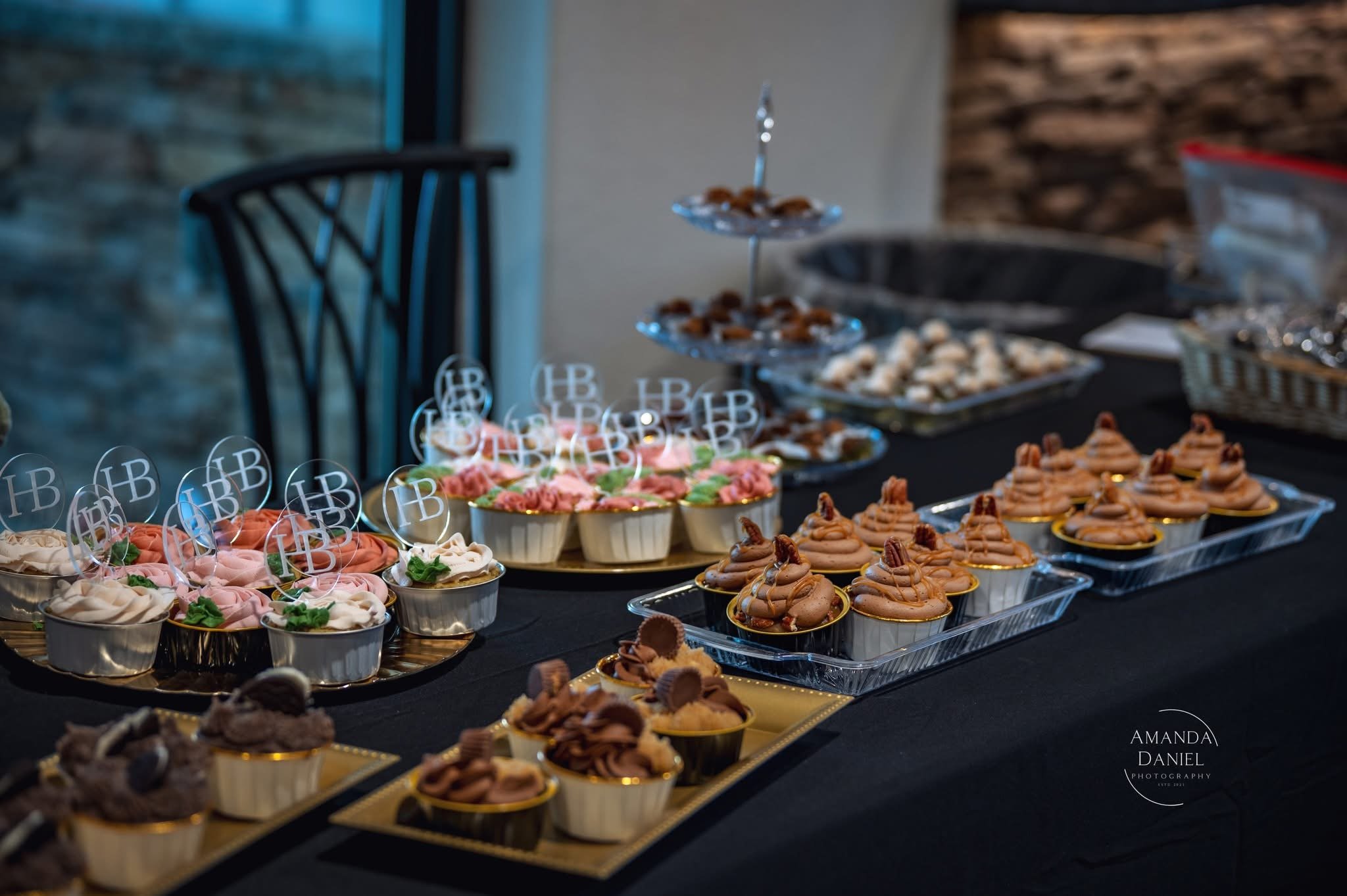 Assorted cupcakes with pink, white, and chocolate frosting decorated with chocolate and cookie toppings, displayed on a black table for a celebration or event.
