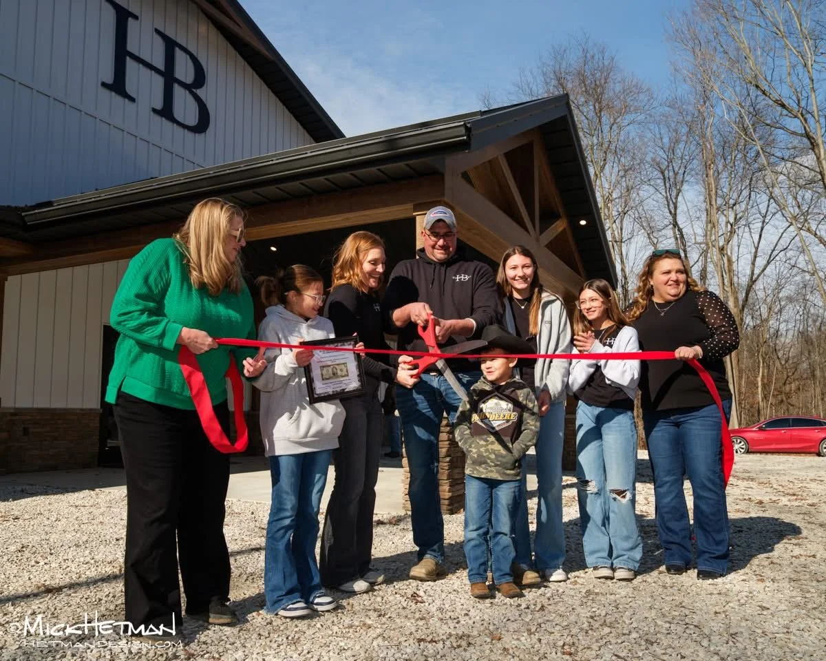 A group of people at a ribbon-cutting ceremony outside a building with the letters 'HB' on it, with some holding a pair of scissors, standing on gravel with trees and a red car in the background.
