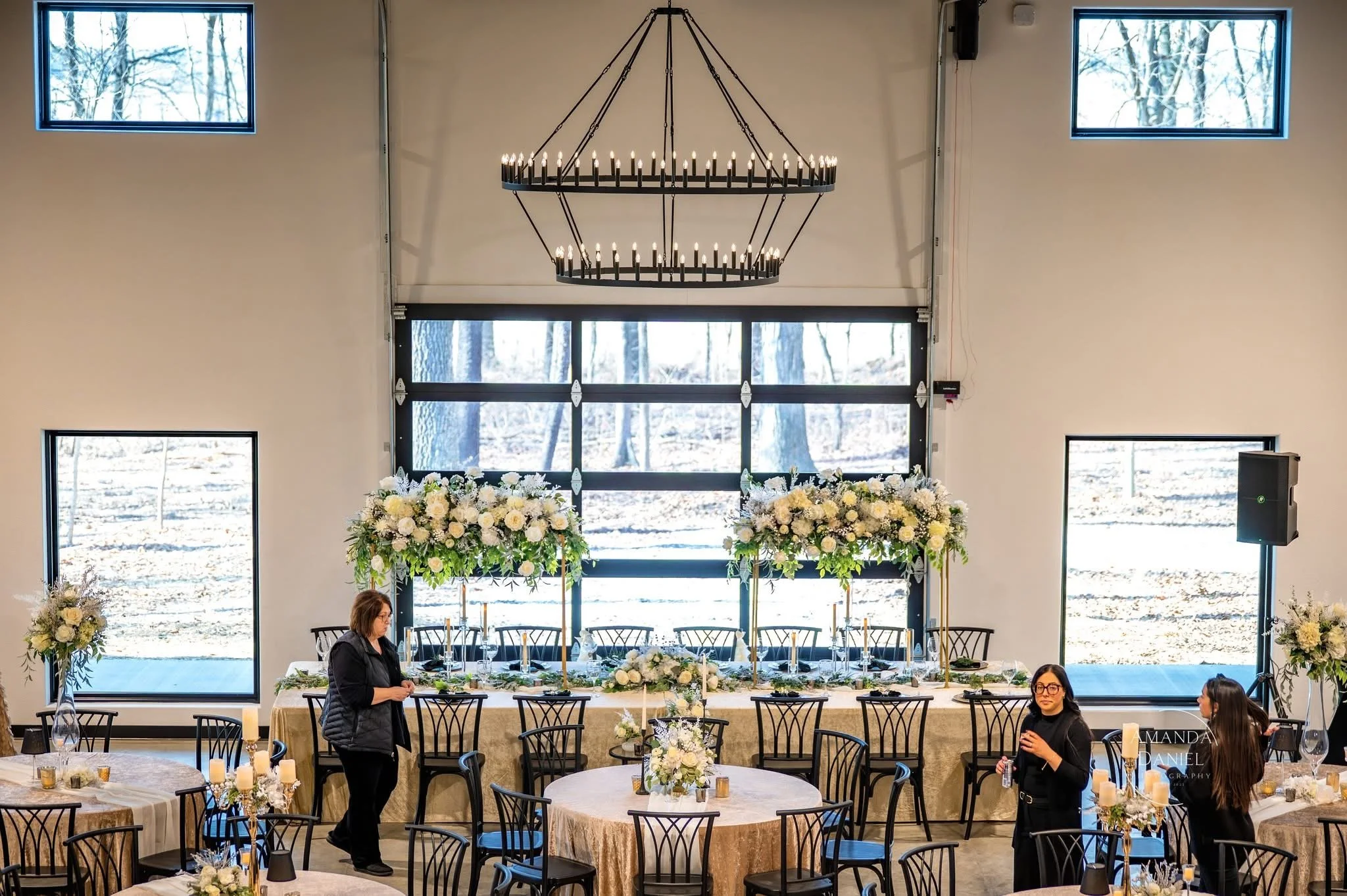 Indoor banquet hall decorated with cream-colored tablecloths, floral centerpieces, and large floral arrangements on the main table. The room has large windows, a high ceiling, and a black chandelier hanging overhead. There are three women, two standi