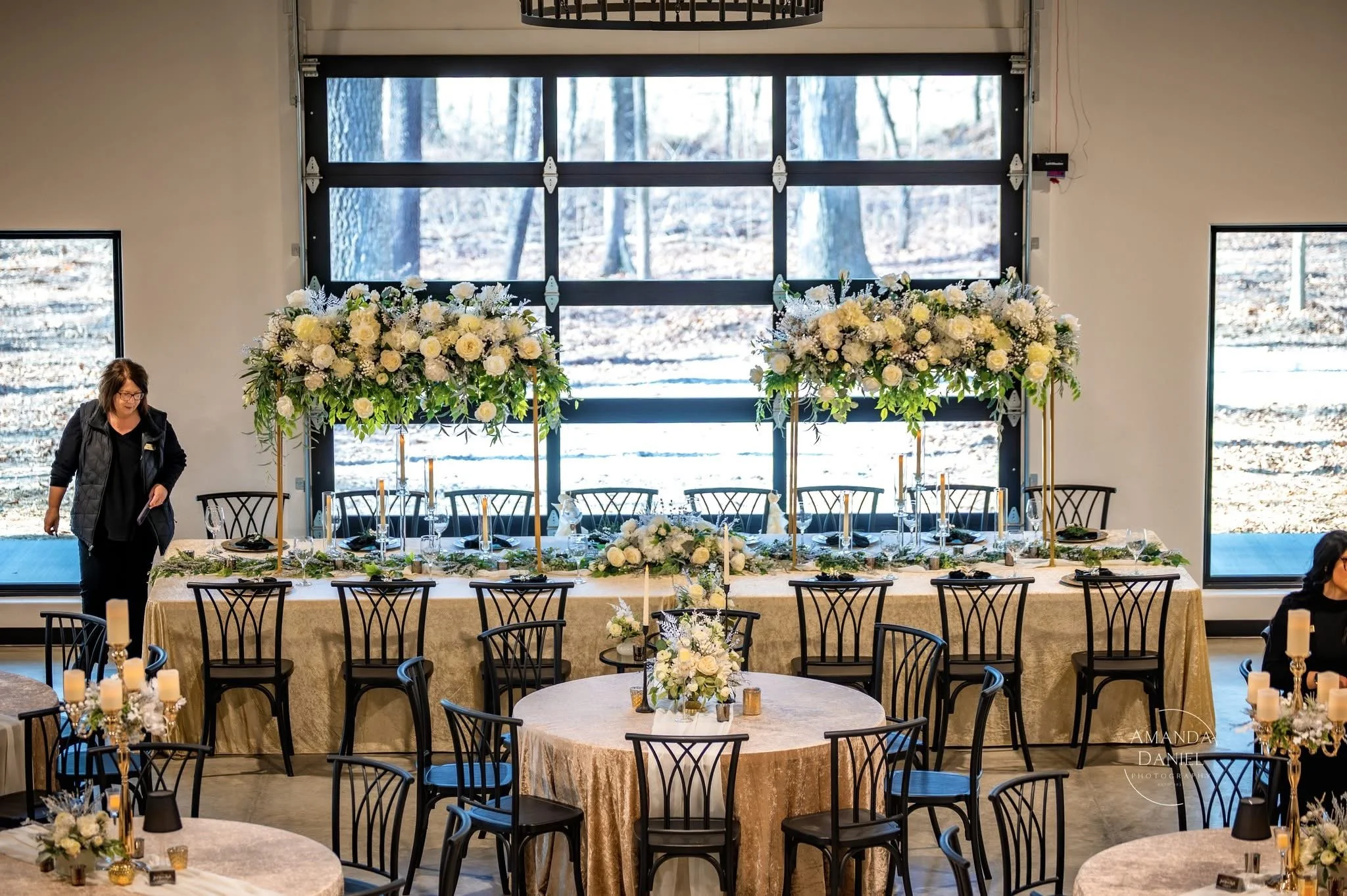 Event space decorated with elegant table settings and floral arrangements. A large rectangular table with tall floral centerpieces is in front of a large window showing a snowy outdoor scene. Round tables with candles and floral decorations are arran