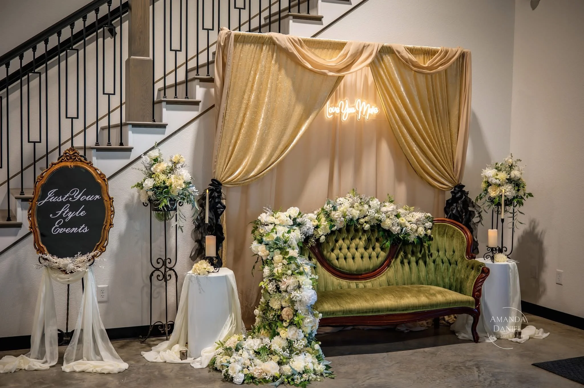 Elegant event setup featuring a vintage green velvet sofa with floral arrangement, a gold draped backdrop with a neon sign that says 'Love You More,' floral stands, candles, and a chalkboard sign that reads 'Just Your Style Events' on a stand.