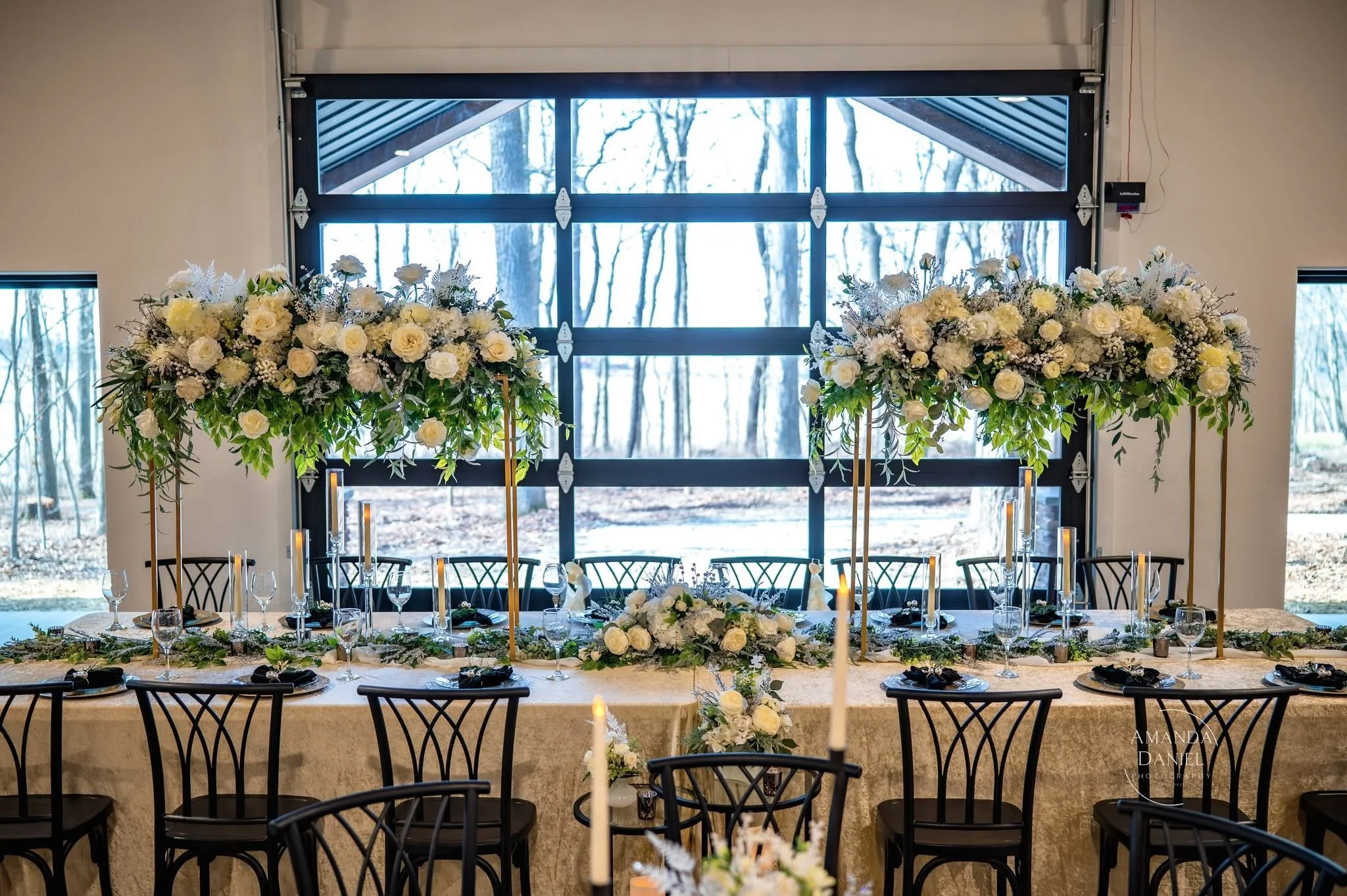 Elegant banquet table decorated with white flowers and tall candle holders in front of large windows with a view of a wooded area.