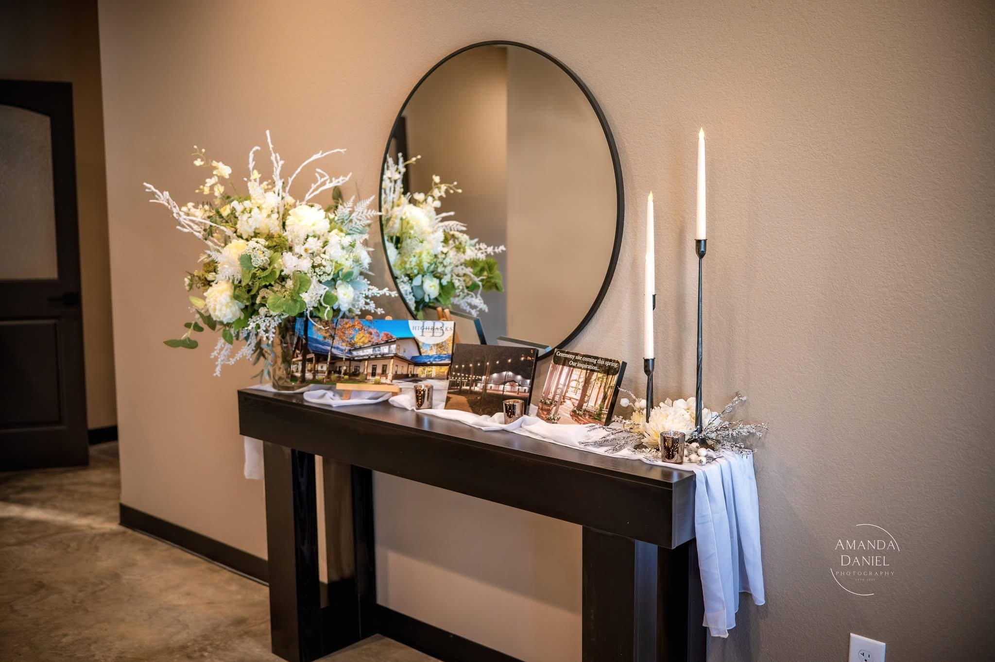Wedding or special event welcome table with white floral arrangement, framed photos, candles, and tall candles on a dark wooden table against a beige wall.