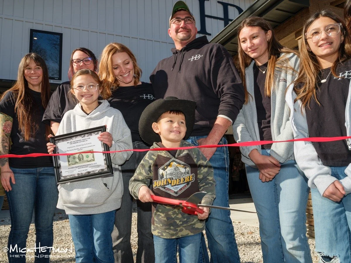 Group of people, including children and adults, gathered outside for a ribbon-cutting ceremony. Some are smiling, and a few are holding framed certificates. A young boy in cowboy attire is holding a pair of scissors, ready to cut the red ribbon.
