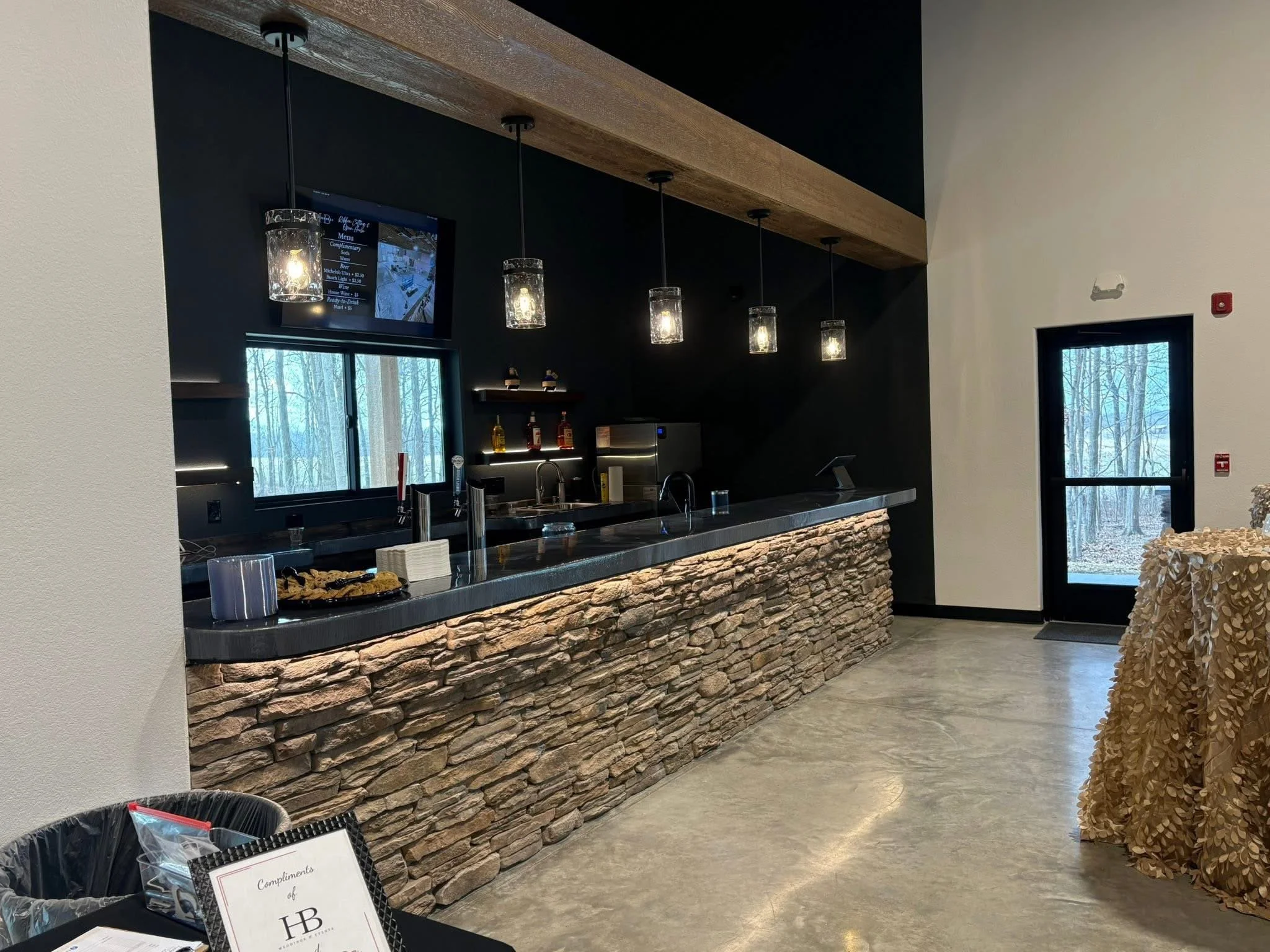 Empty bar area with stone front, black countertop, hanging pendant lights, small window, and a TV screen on the wall. There are bottles on shelf and various items on the bar countertop, with a door to the outdoors at the back.