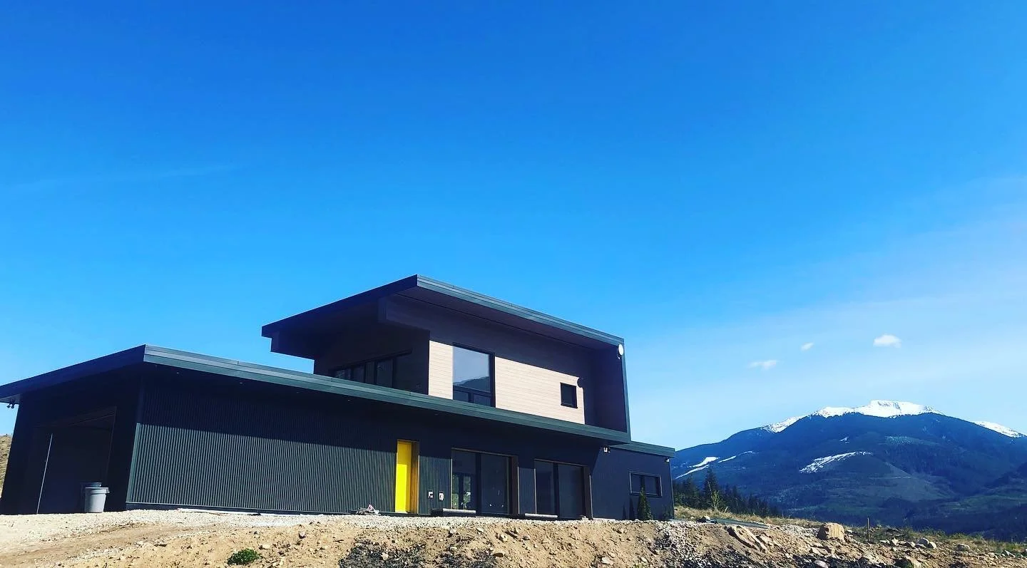 Modern black and wood-clad home with a bold yellow door, set against a backdrop of mountains and clear blue skies in the Sea to Sky Corridor