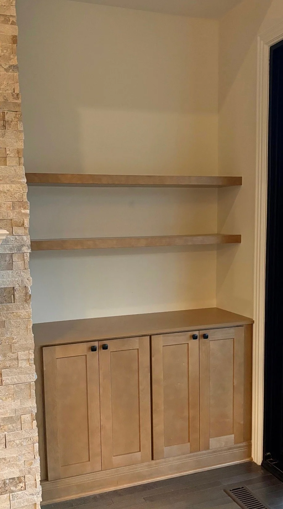 Empty built-in wooden shelving unit with three open shelves and a cabinet with four doors, set against a wall in a room with stone and wall paneling.