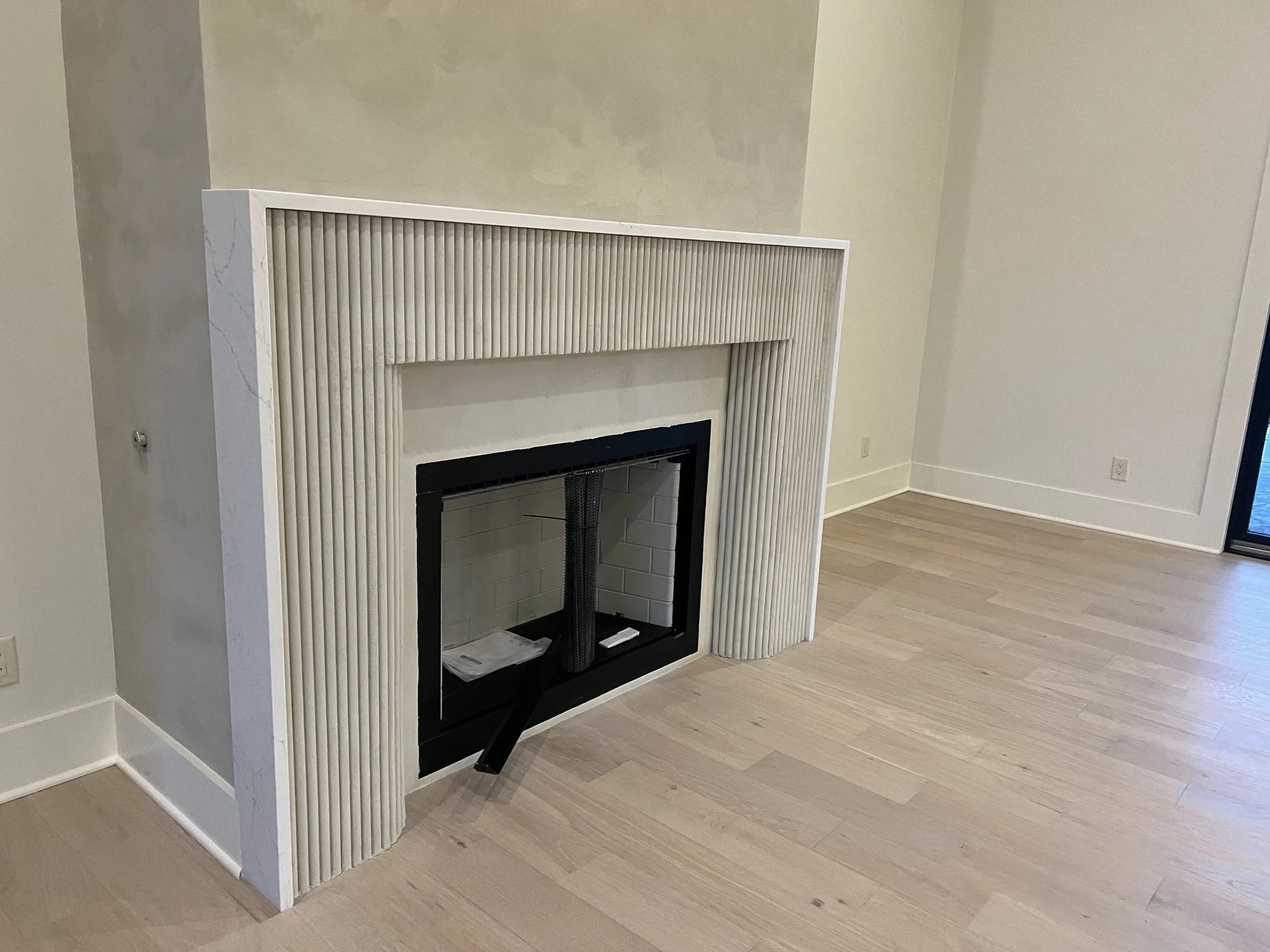 Modern fireplace with beige, textured, ribbed frame and black hearth in a living room with light wood flooring and white walls.