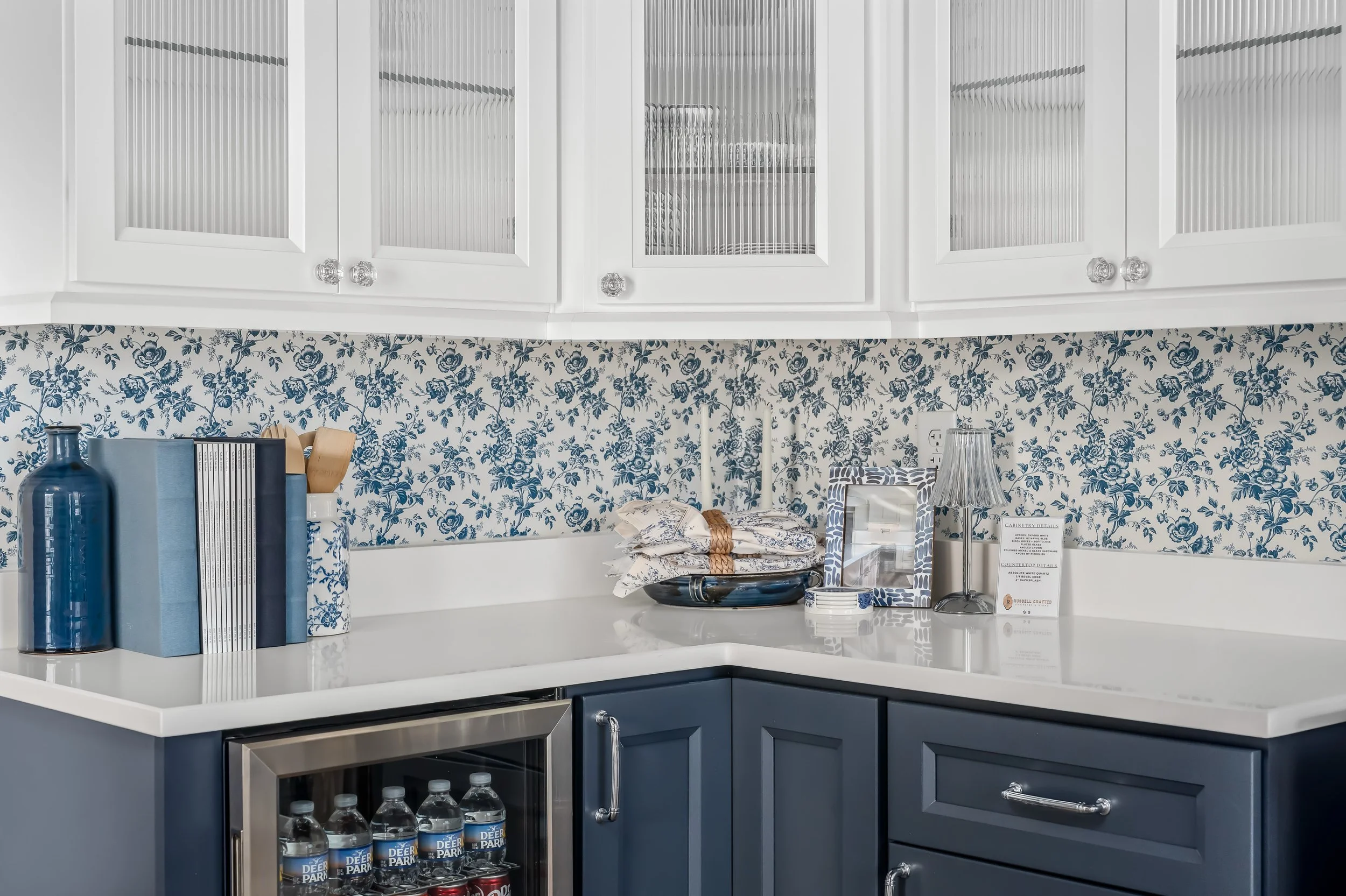 Kitchen corner with white upper cabinets with glass doors, blue and white floral wallpaper, blue lower cabinets, a white countertop with bottles of water, books, and decorative items.