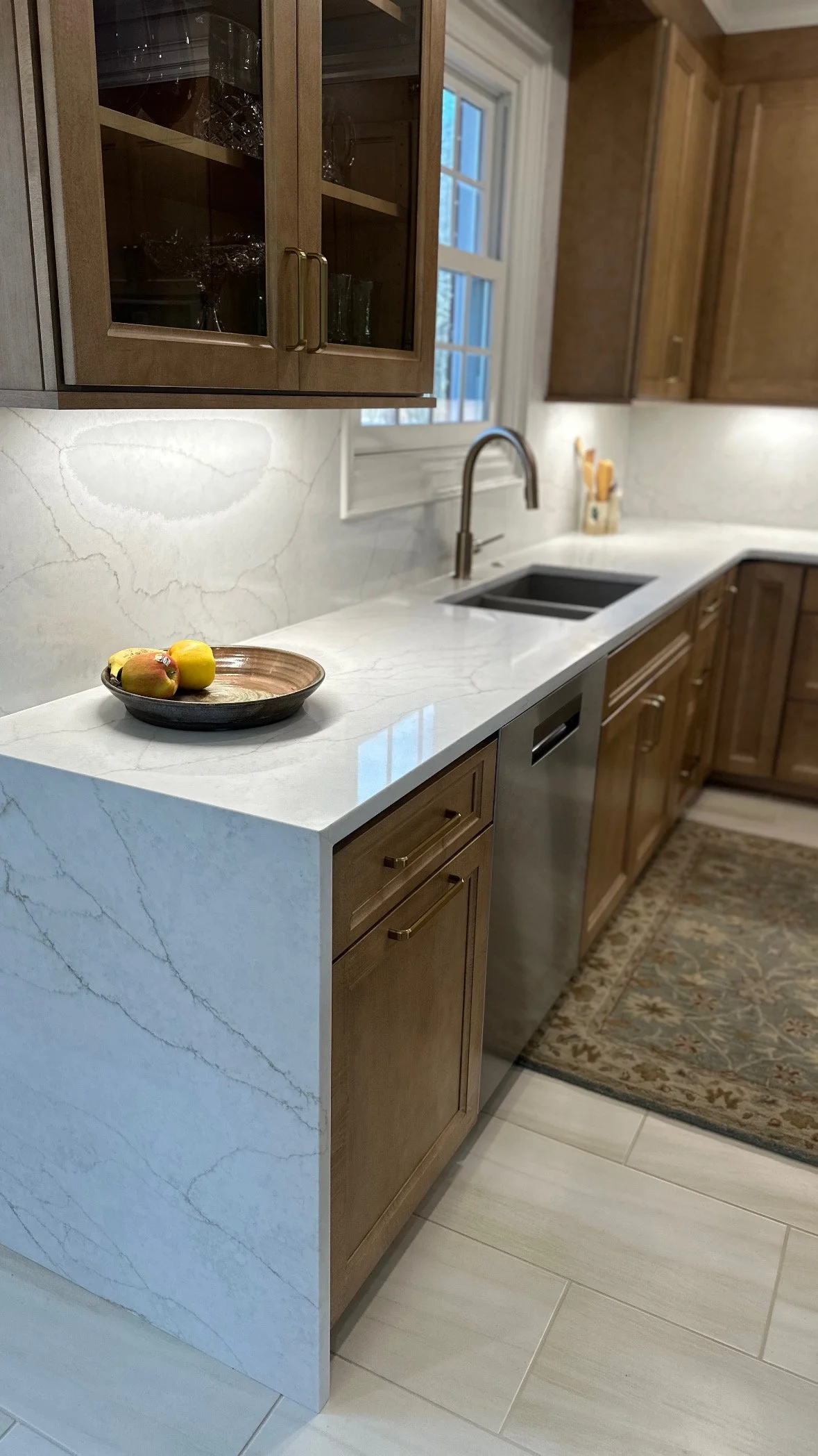 Kitchen with white marble countertop, wooden cabinetry, stainless steel dishwasher, and a black sink. A brown wooden dish with apples is on the counter near the corner.