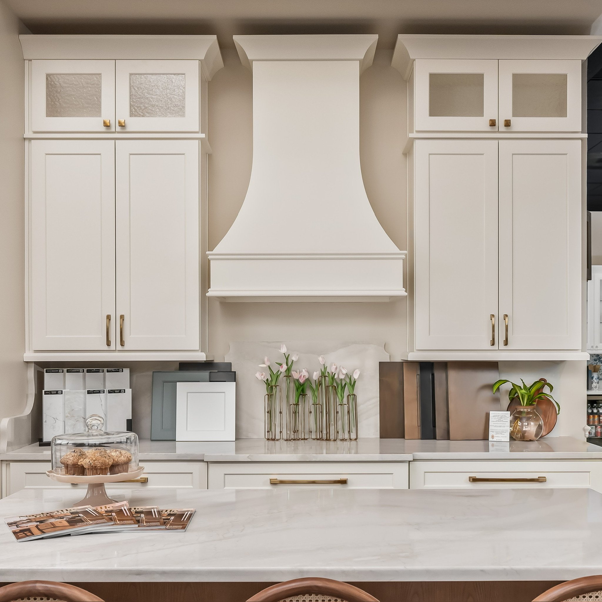 White kitchen cabinets with glass doors, a marble countertop, a cake stand with baked goods, floral arrangements, books, and a potted plant.