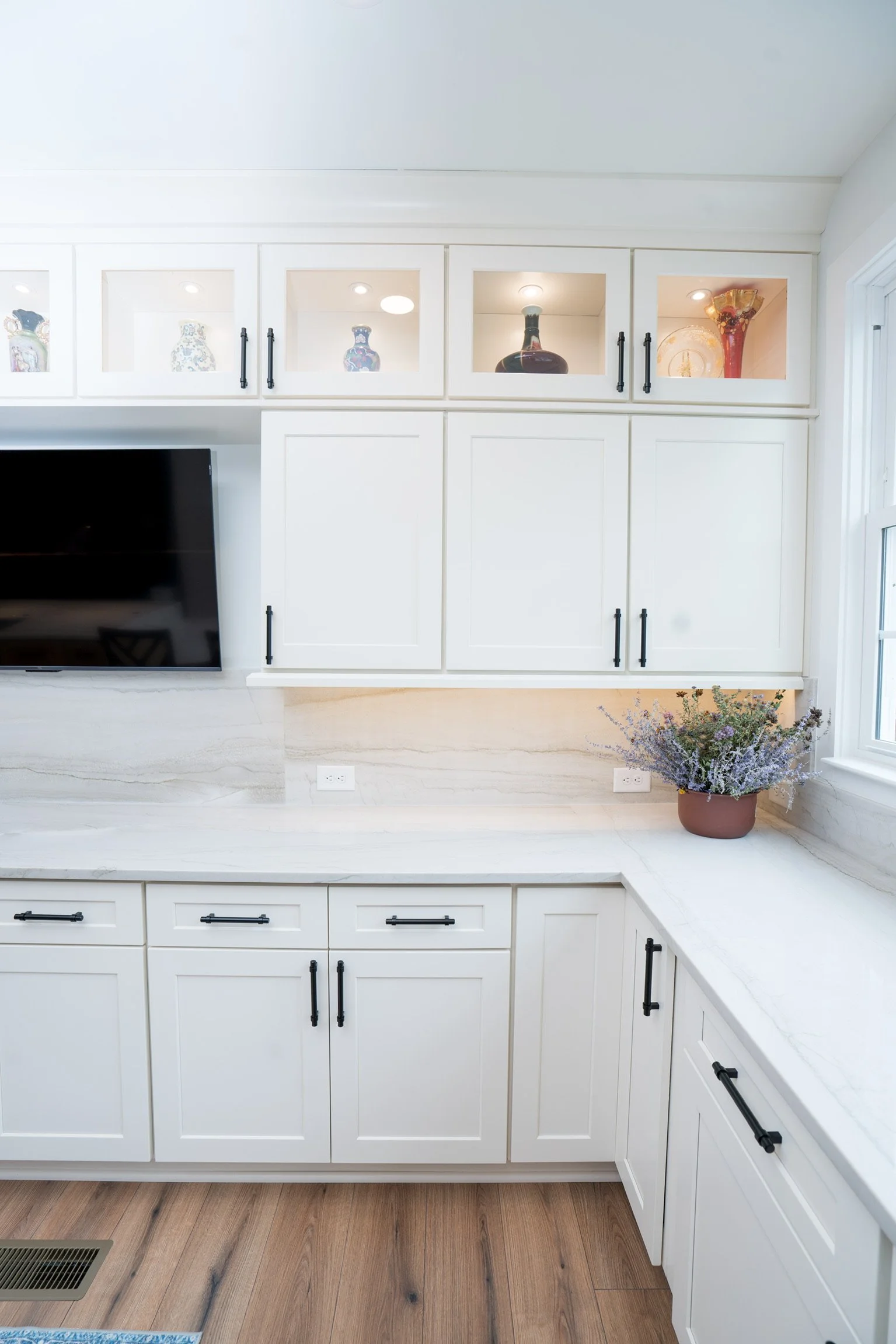 White kitchen cabinets with black handles, marble countertops, a potted lavender plant, a flat-screen TV on the left, and glass-front upper cabinets displaying decorative vases and a large bottle.