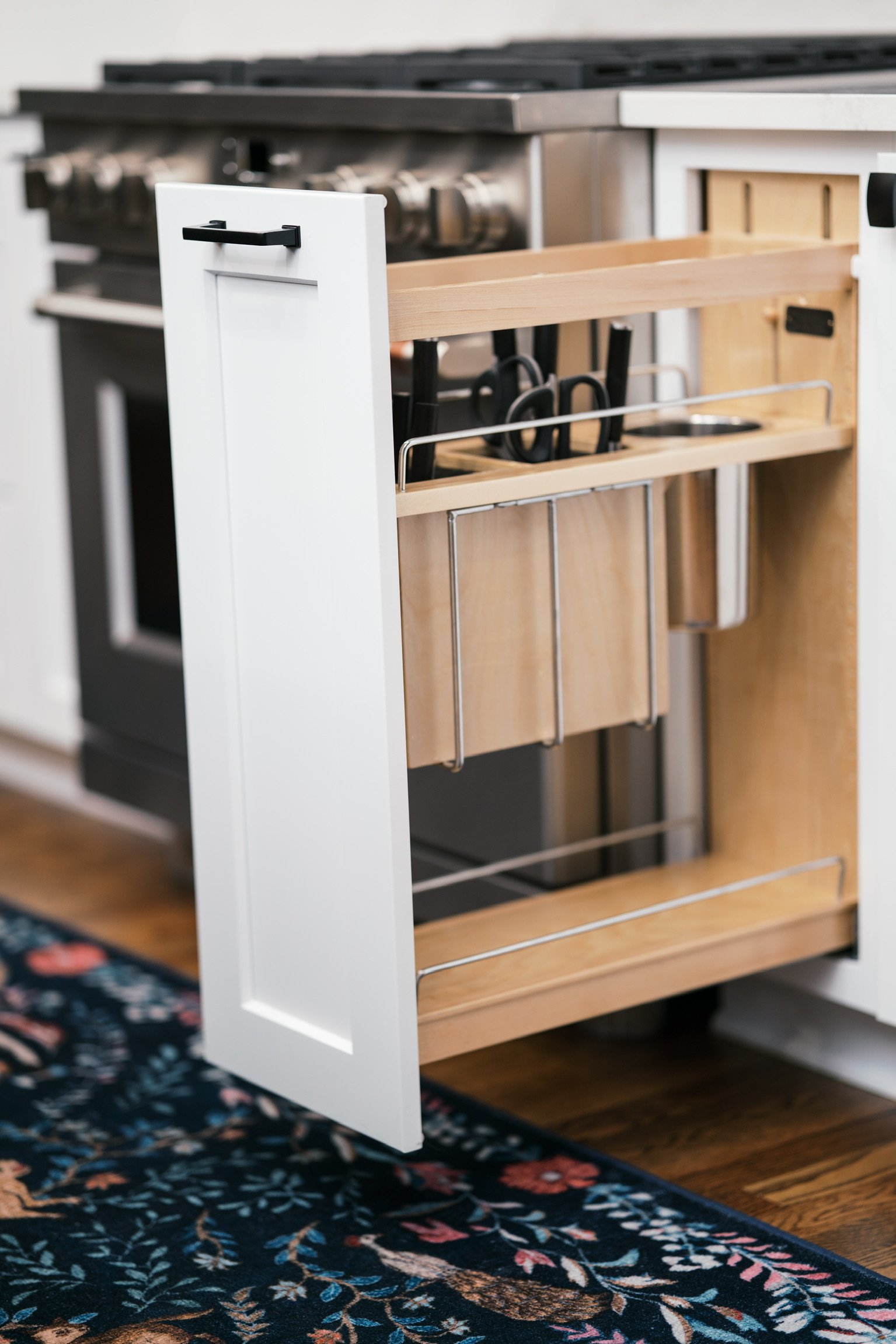 Open kitchen cabinet with a wooden utensil tray holding scissors, placed in front of a stainless steel stove.
