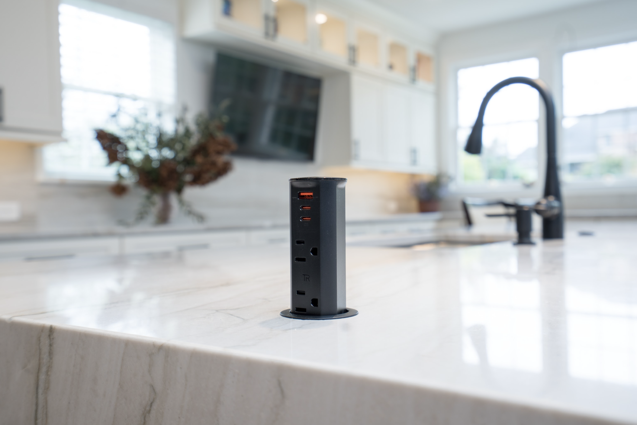 A black power strip test socket with multiple outlets and USB ports is mounted on a marble kitchen countertop, with a modern kitchen in the background featuring white cabinets, a stainless steel faucet, and large windows.