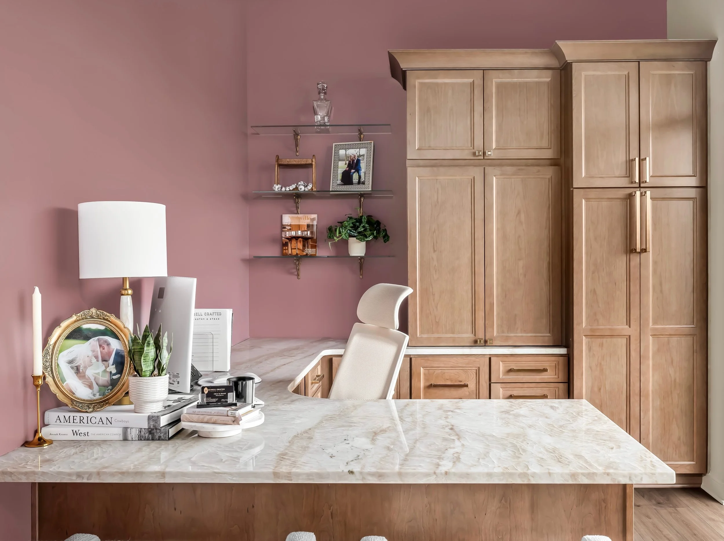Home office with pink wall, marble desk, beige chair, wooden cabinetry, glass shelves with pictures and decor, laptop, books, and framed wedding photo.