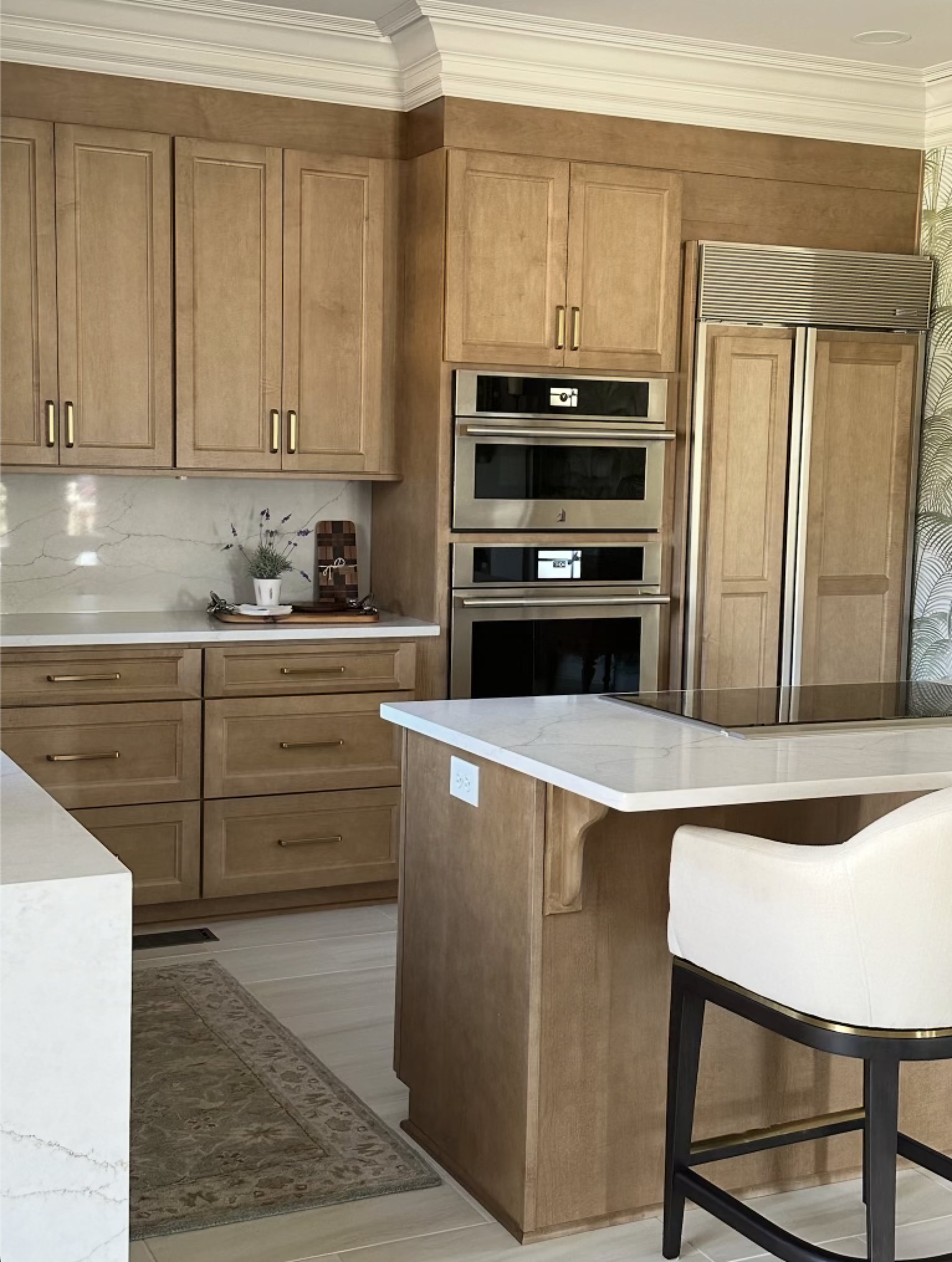 Kitchen with wooden cabinetry, built-in stainless steel double ovens, a microwave, large refrigerator, white marble countertops, a small potted plant, and a barstool.