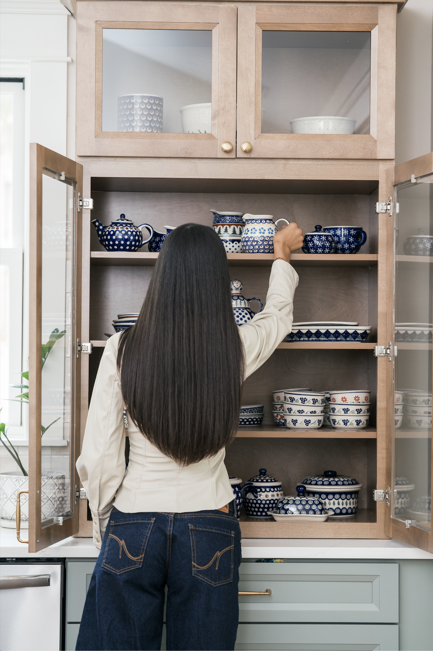A woman with long dark hair reaching into a wooden cabinet filled with blue and white patterned dishware.