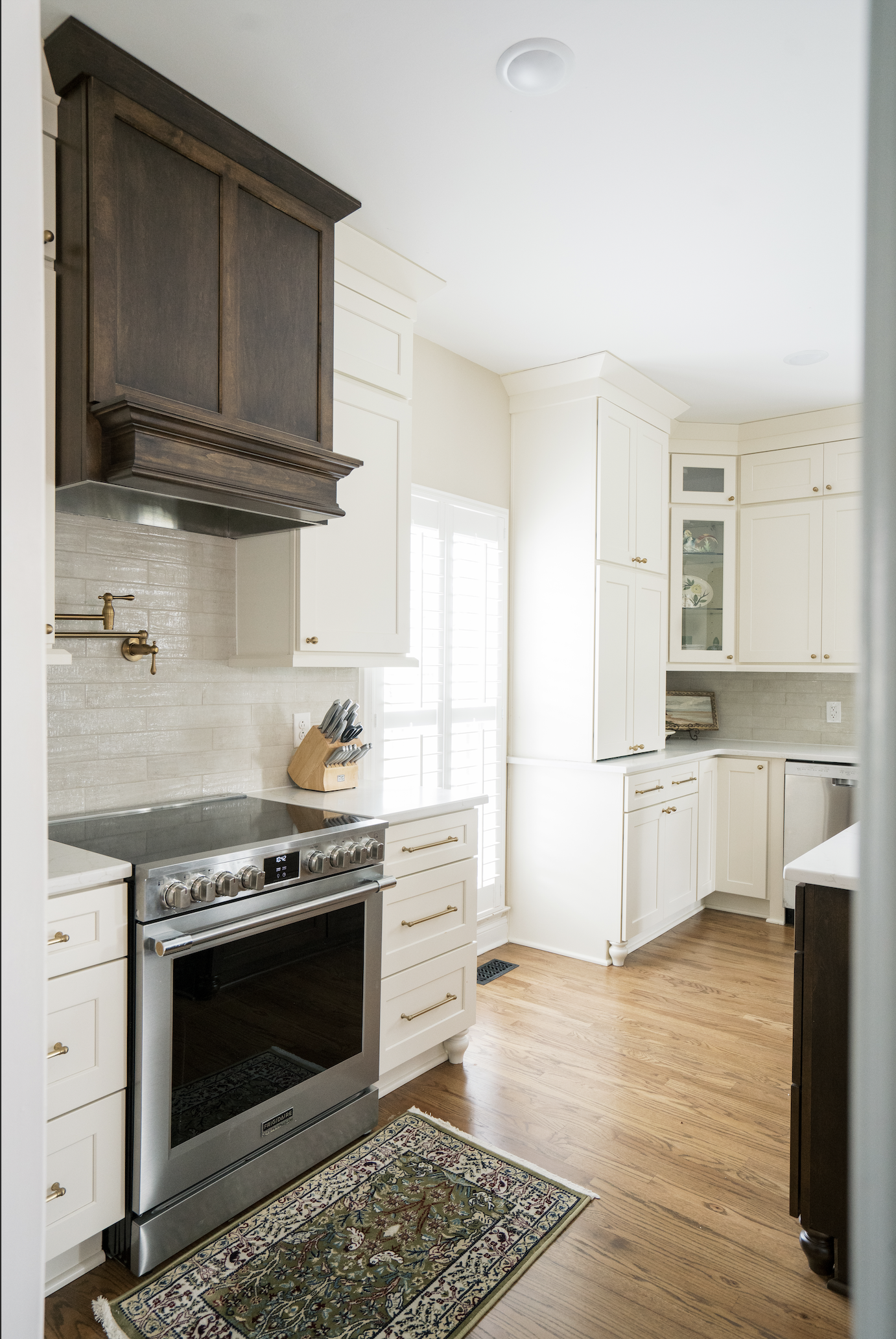 Bright kitchen with white cabinets, a stainless steel oven, a knife block, a window with white shutters, and a patterned rug on hardwood floor.