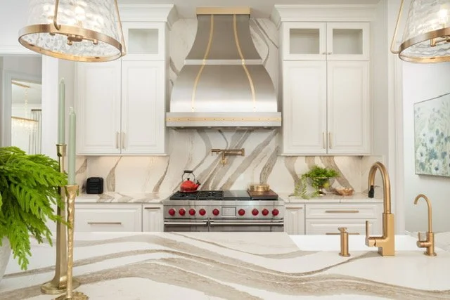 Modern kitchen with white cabinets, marble backsplash, red and black stove, gold fixtures, and decorative lighting.