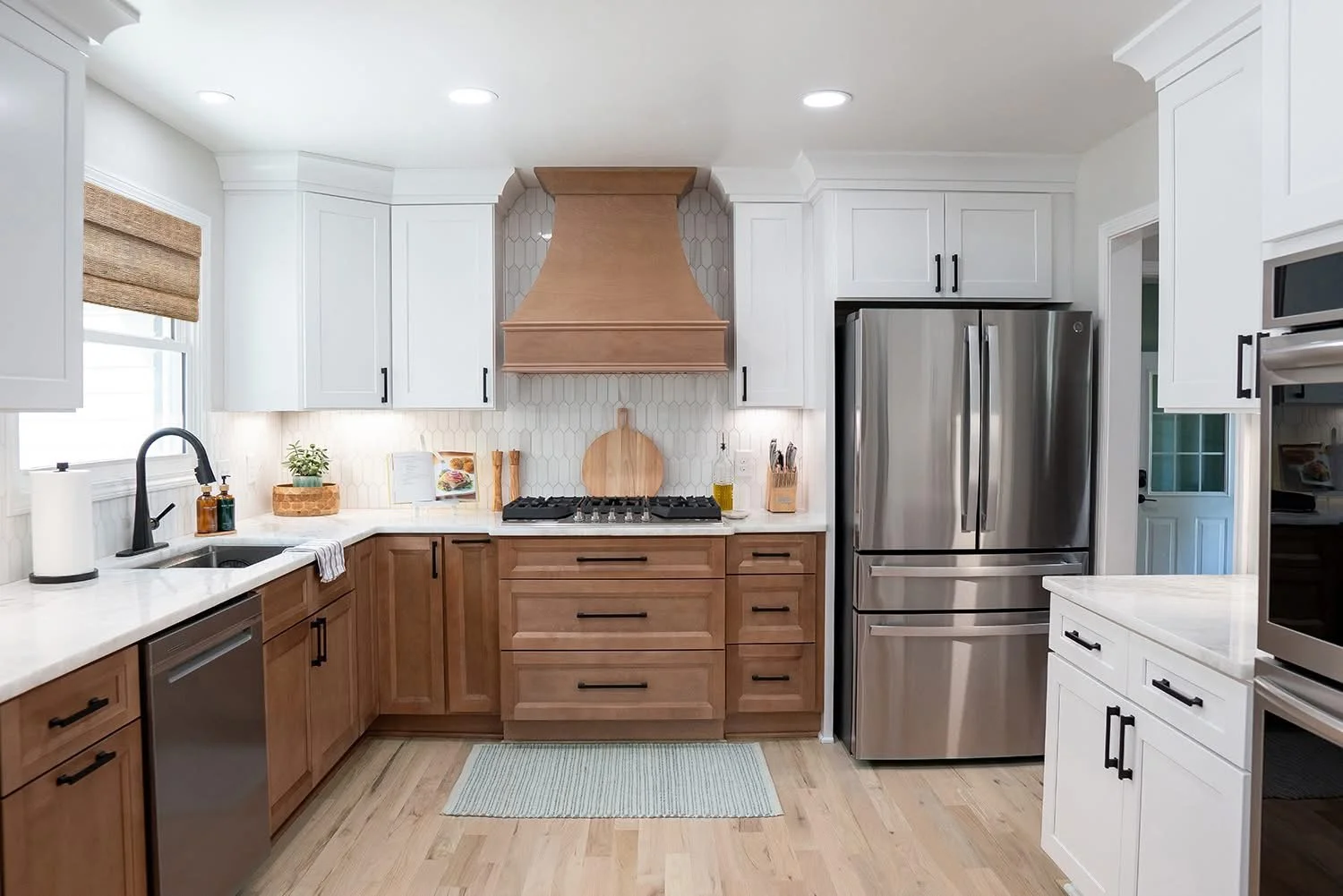Modern kitchen with wood and white cabinetry, stainless steel refrigerator, gas stove with wooden range hood, white marble countertops, small window with woven shade, and hardwood floors.