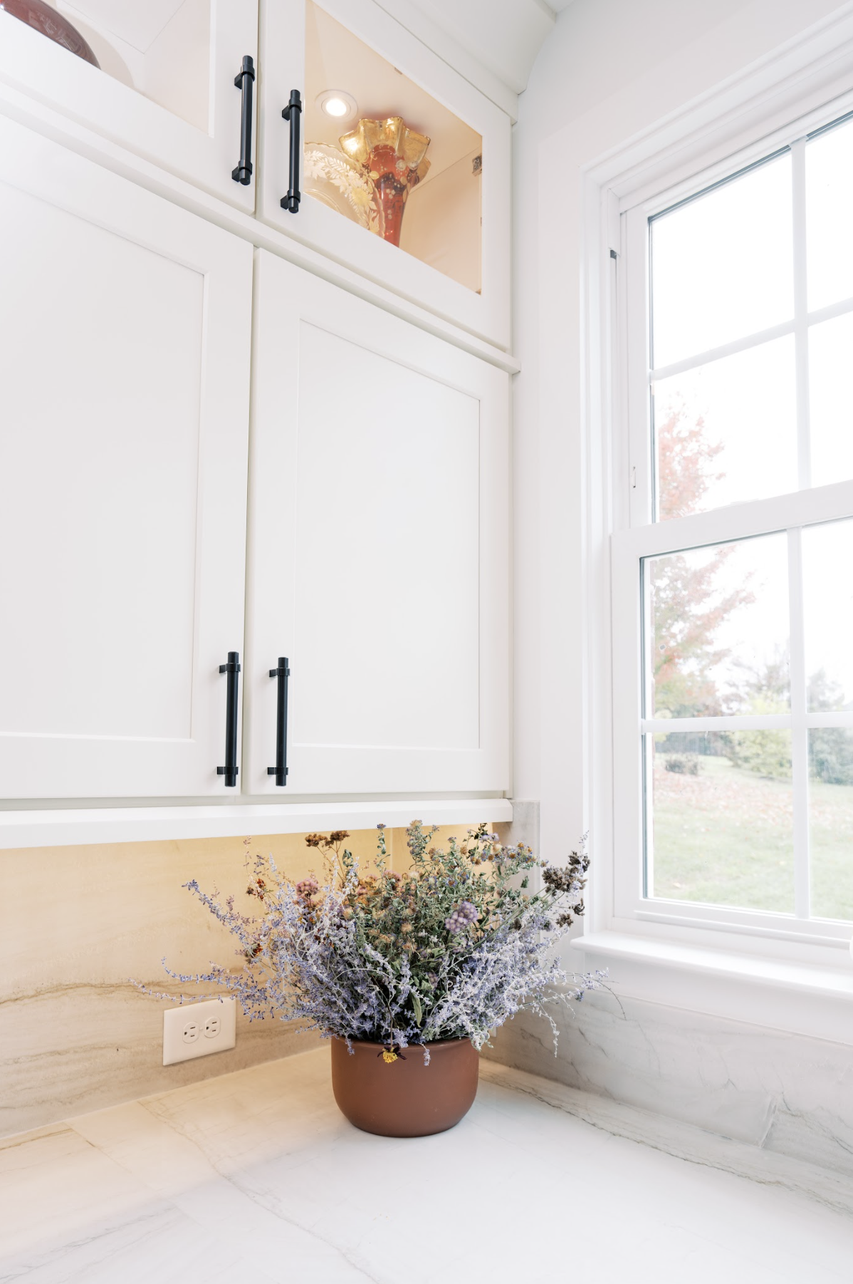 A cozy corner of a kitchen with a vase of purple and green flowers on a white marble countertop, a large window with white framing showing trees outside, and a white cabinet with black handles and a decorative ceramic vase inside.