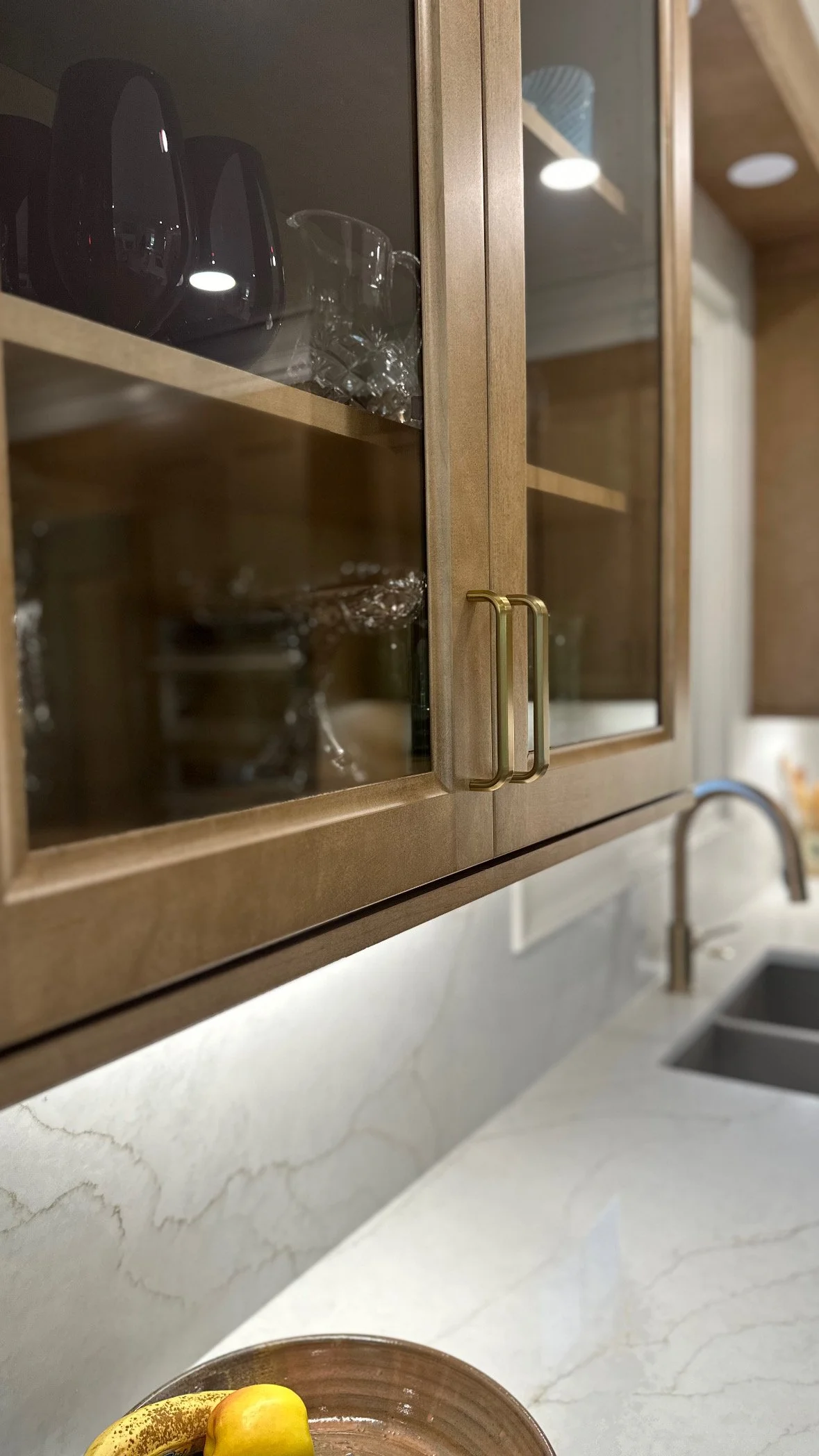 Wooden kitchen cabinet with glass doors and brass handles, white marble countertop with a sink, and a bowl of bananas and yellow fruit in the foreground.