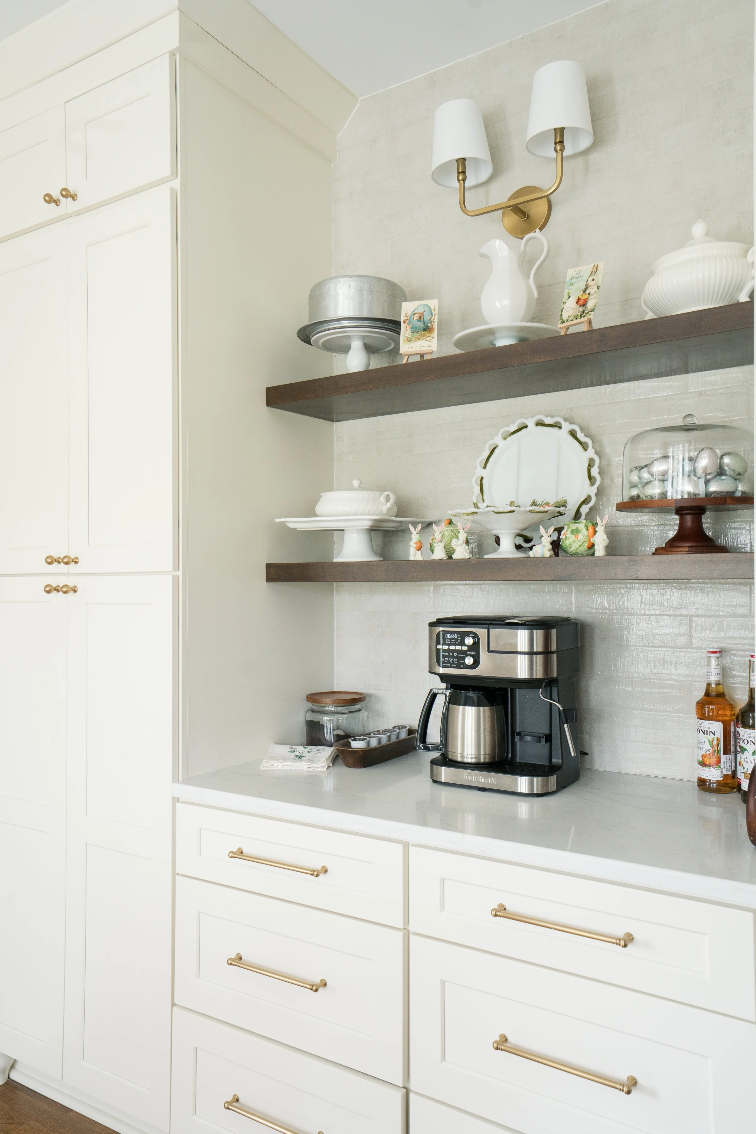 Kitchen countertop with a coffee maker, jars, and decorative items. Two wooden shelves above hold dishes, teapots, and decorative figurines. White cabinets with gold handles on the left.